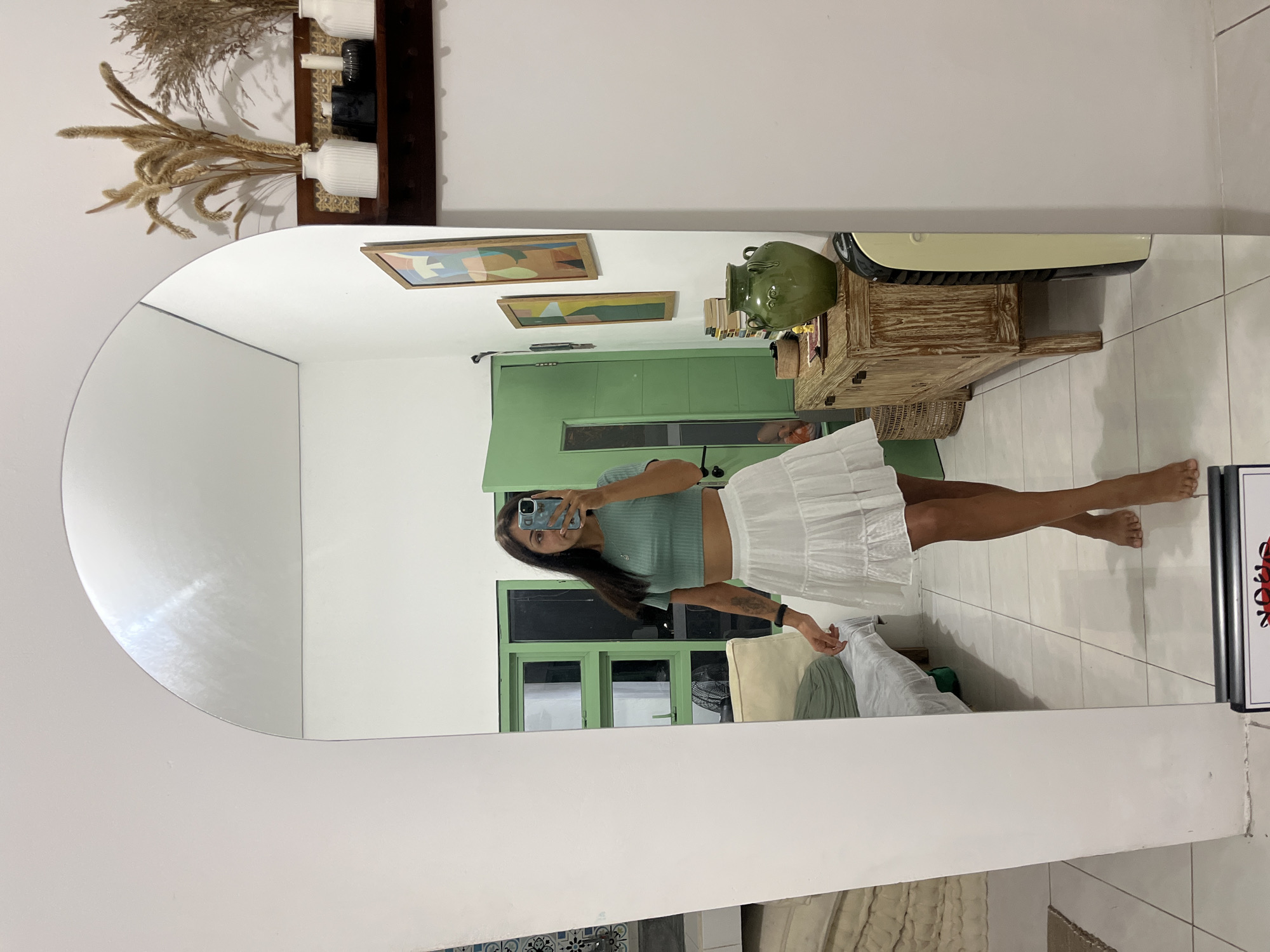 A full-body mirror selfie captures a young woman posing indoors in Kuta Selatan, Indonesia. The image is oriented sideways (rotated 90 degrees clockwise from vertical). The woman, with long dark hair, is wearing a light teal ribbed short-sleeved crop top and a white tiered ruffled mini-skirt. She is barefoot and stands on a light-colored tiled floor. Her left arm has visible tattoos. She holds a light blue smartphone with a patterned case up to her face, obscuring her expression as she takes the photograph.
The reflection in the large, arch-topped mirror reveals the interior of a room with white walls and a light-colored tiled floor, showcasing a modern, relaxed aesthetic. In the background, a prominent green-painted cabinet unit or door with dark glass panels is visible. Above this unit, two framed abstract artworks with geometric shapes and soft colors hang on the wall. To the right, a rustic wooden console table holds a large green ceramic urn, a stack of books, and other small decorative items. An air conditioning unit, cream and black, is mounted on the wall above the table.
Part of a bed with light-colored bedding and pillows is visible in the lower right of the reflection. Above the mirror's arch, a wall-mounted shelf displays dried botanical arrangements, including feathery grasses like pampas, alongside white and black decorative vases. On the far right of the reflection, a partial view of a white sign with black text reading "HOOK" and some red graphical elements is visible. The overall setting suggests a comfortable and stylish living space.