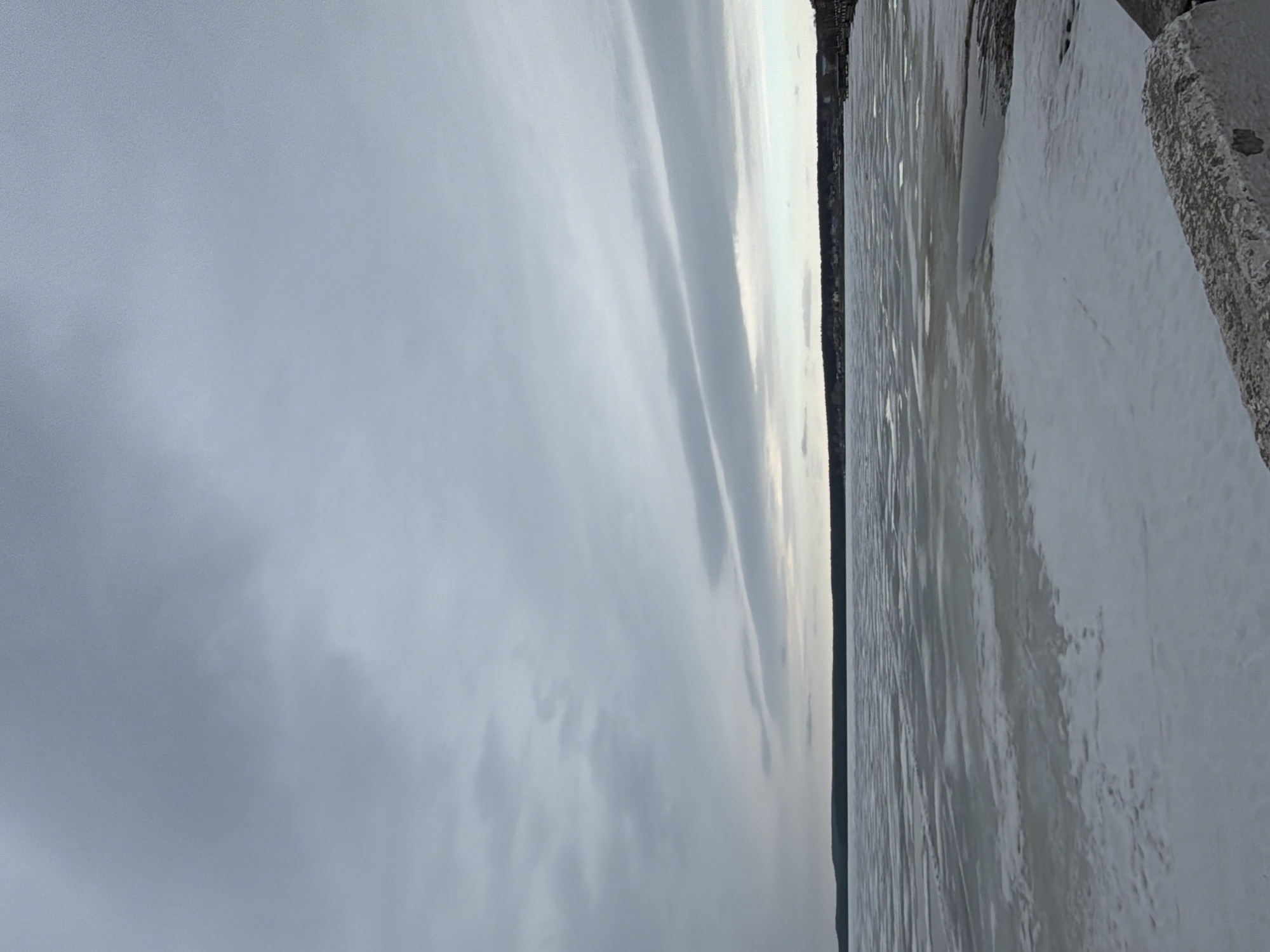 The image depicts a view of a cloudy sky over a body of water, possibly a lake or river, adjacent to a shoreline with building structures. The picture was taken from a slightly elevated position, capturing the water’s surface with ripples or small waves. The shoreline includes a concrete structure, possibly a pier or embankment. Across the water, a faint outline of a town is visible, consisting of houses and buildings. The scene suggests an overcast or wintery day. Given the information, it is likely that this image was captured in the city of Votkinsk, Russia, likely overlooking the Votkinsk Reservoir. The image is rotated 90 degrees clockwise.