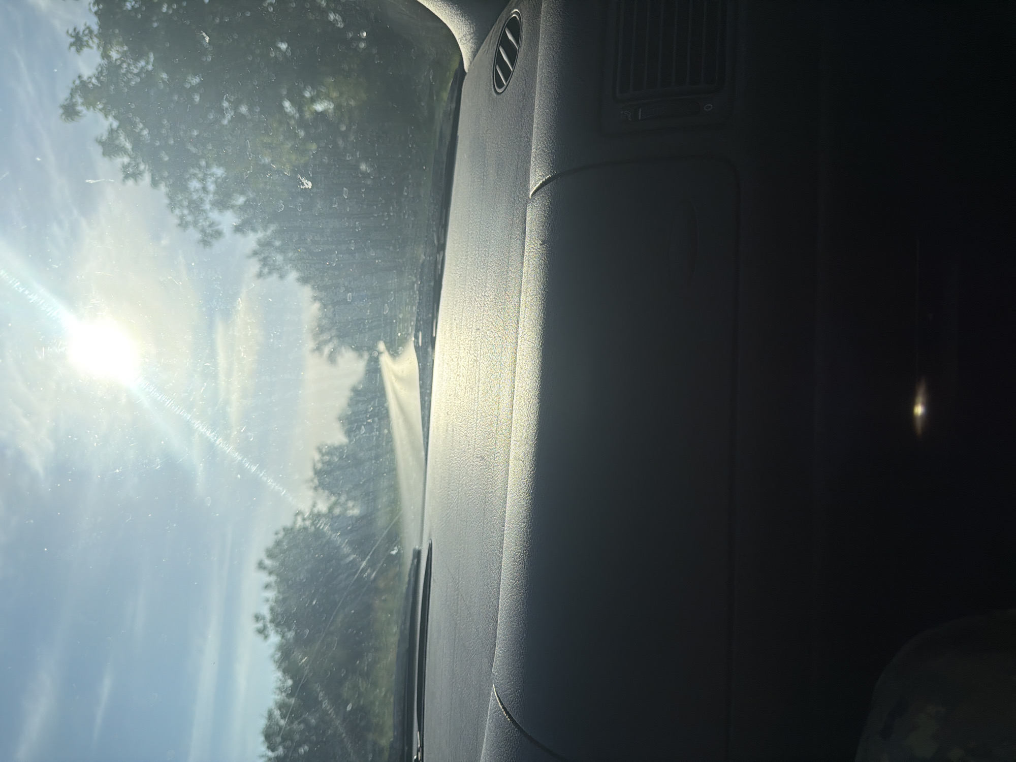 The image shows the interior of a car, specifically the dashboard and windshield. The scene is bright due to sunlight shining through the windshield. Through the windshield, trees and the sky are visible. The dashboard is gray and shows details such as air vents. The location cannot be determined from the image. There are no people visible.