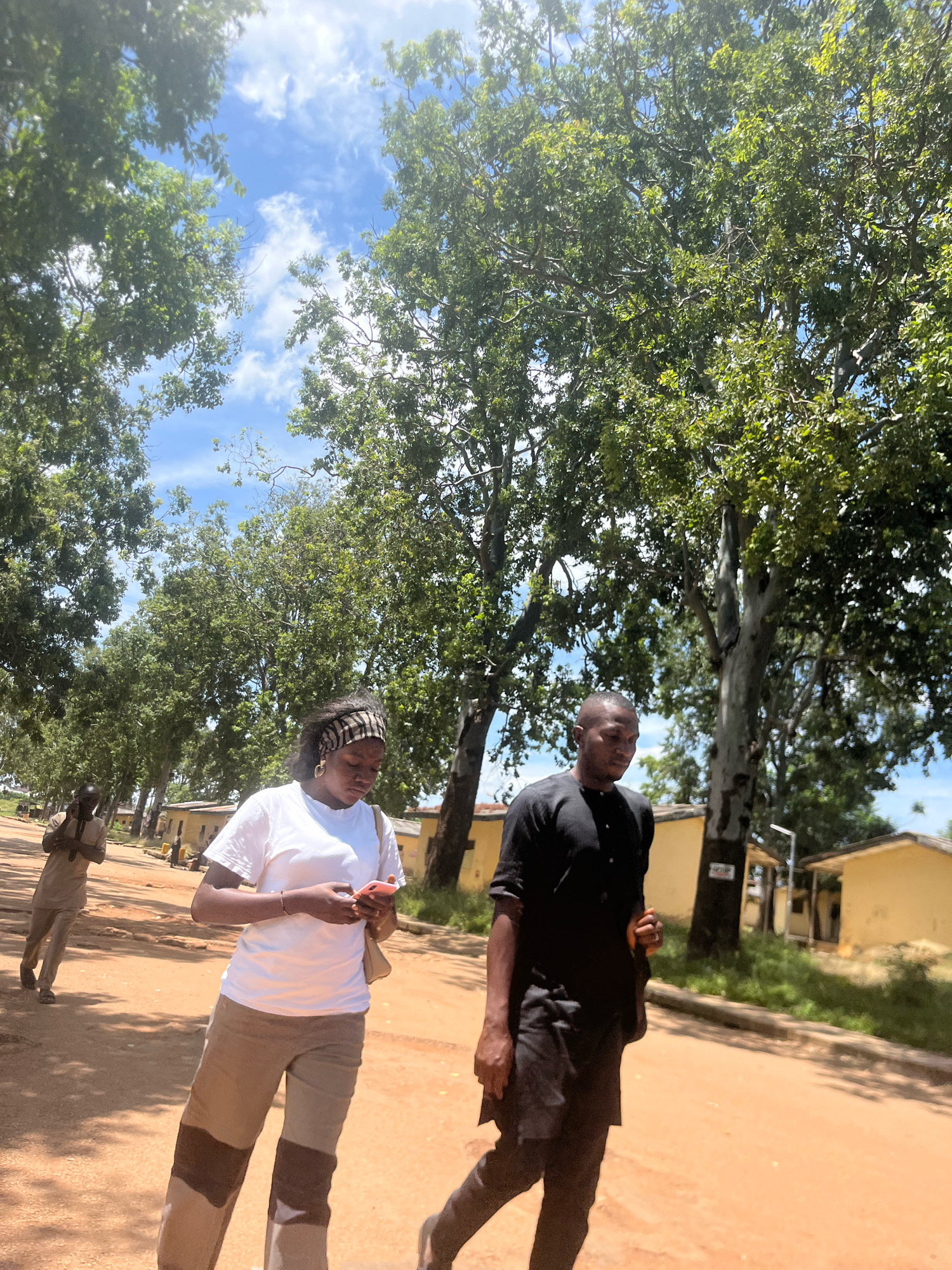 In Rafin Par, Nigeria, two individuals, a woman and a man, are walking towards the camera on a dusty, unpaved road. The woman, in the foreground on the left, wears a white t-shirt and beige pants with brown accents. She is looking down at her pink smartphone, which she holds in her hands. A light-colored bag is slung over her left shoulder. To her right and slightly behind her, the man is dressed in a black, long-sleeved, collared tunic and dark pants. He is walking with his head down and slightly to the left. In the background, on the left side of the road, another person is walking away from the camera, wearing a light-brown outfit. Along the right side of the road, there are several small, single-story yellow buildings with pitched roofs, and mature trees with lush green foliage line the road on both sides, creating a canopy. The sky is blue with scattered white clouds. The overall scene suggests a bright, sunny day in a rural or semi-urban setting.