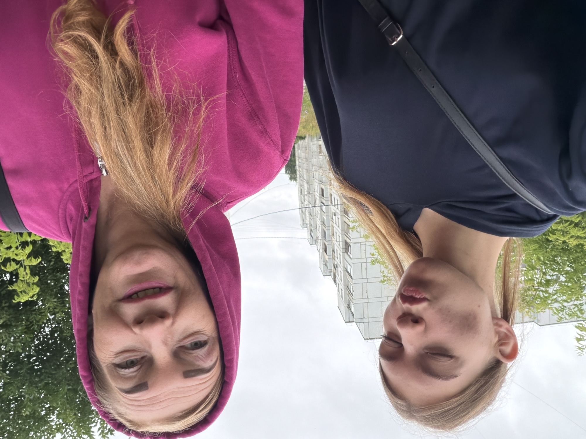 Two individuals are captured in a close-up, outdoor selfie. The person on the left, a younger female with blonde hair pulled into pigtails, wears a dark navy hooded sweatshirt and a black crossbody bag. Her eyes are closed, and her lips are pursed. She has dark marks on her cheeks. The person on the right, an older female with blonde hair, is wearing a bright pink hooded sweatshirt. She has prominent eyebrows and is smiling with her mouth slightly open. They are positioned in front of a multi-story apartment building and green trees under a cloudy sky. The image appears to be taken in the city of Chaikovsky, Russia, given the architectural style of the building and the overall setting.