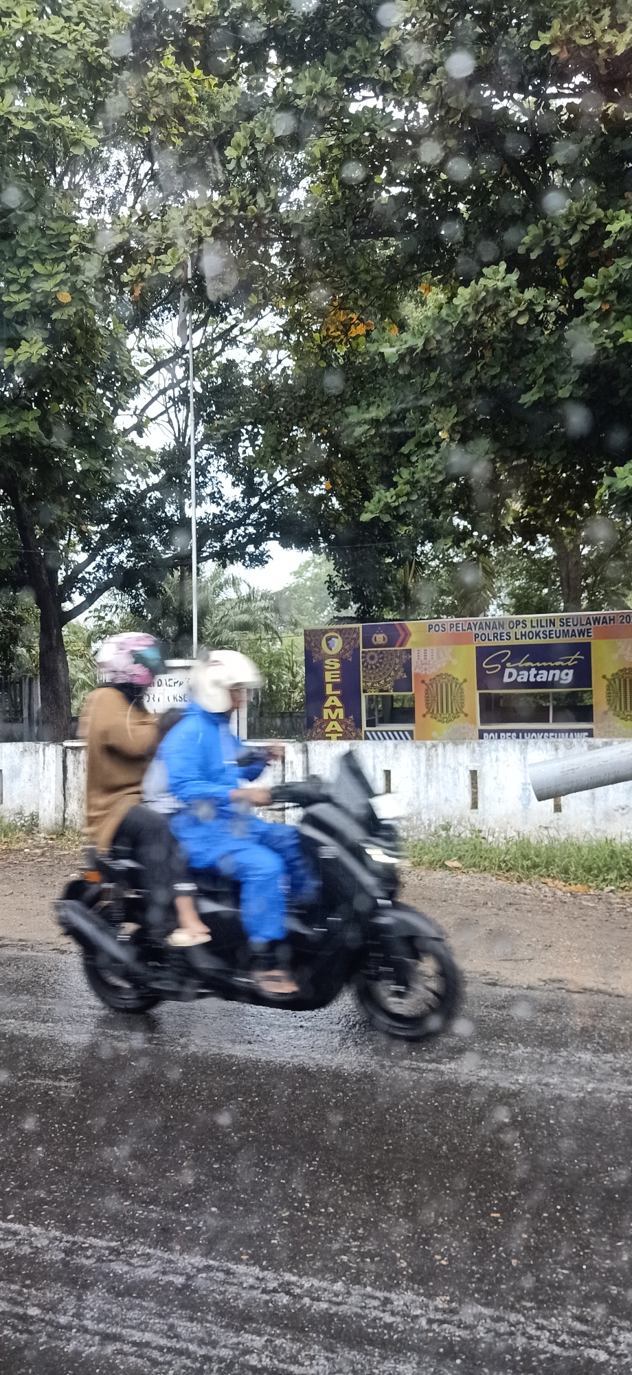 Two individuals are riding a black scooter on a wet road. The person in front is wearing a brown long-sleeved shirt and black pants, with a pink and black helmet. The person behind is wearing a blue jumpsuit and a white helmet. They are riding past a white wall with a sign that reads "POS PELAYANAN OPS LILIN SEULAWAH 2023" and "SELAMAT DATANG POLRES LHOKSEUMAWE". Trees are visible in the background. The scene appears to be in Palohlada, Indonesia, based on the mention of Lhokseumawe. The road is wet, suggesting it has recently rained.