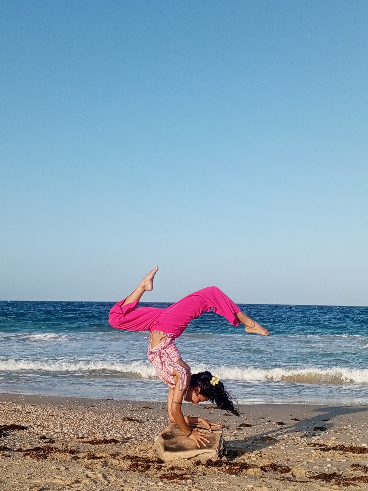A woman is doing a yoga handstand on a rock at a beach. She is wearing pink pants and a patterned top. The ocean is behind her with waves coming to shore. The sky is blue. The beach is sandy with seaweed scattered around. The location appears to be a beach near Porlamar, Venezuela.
