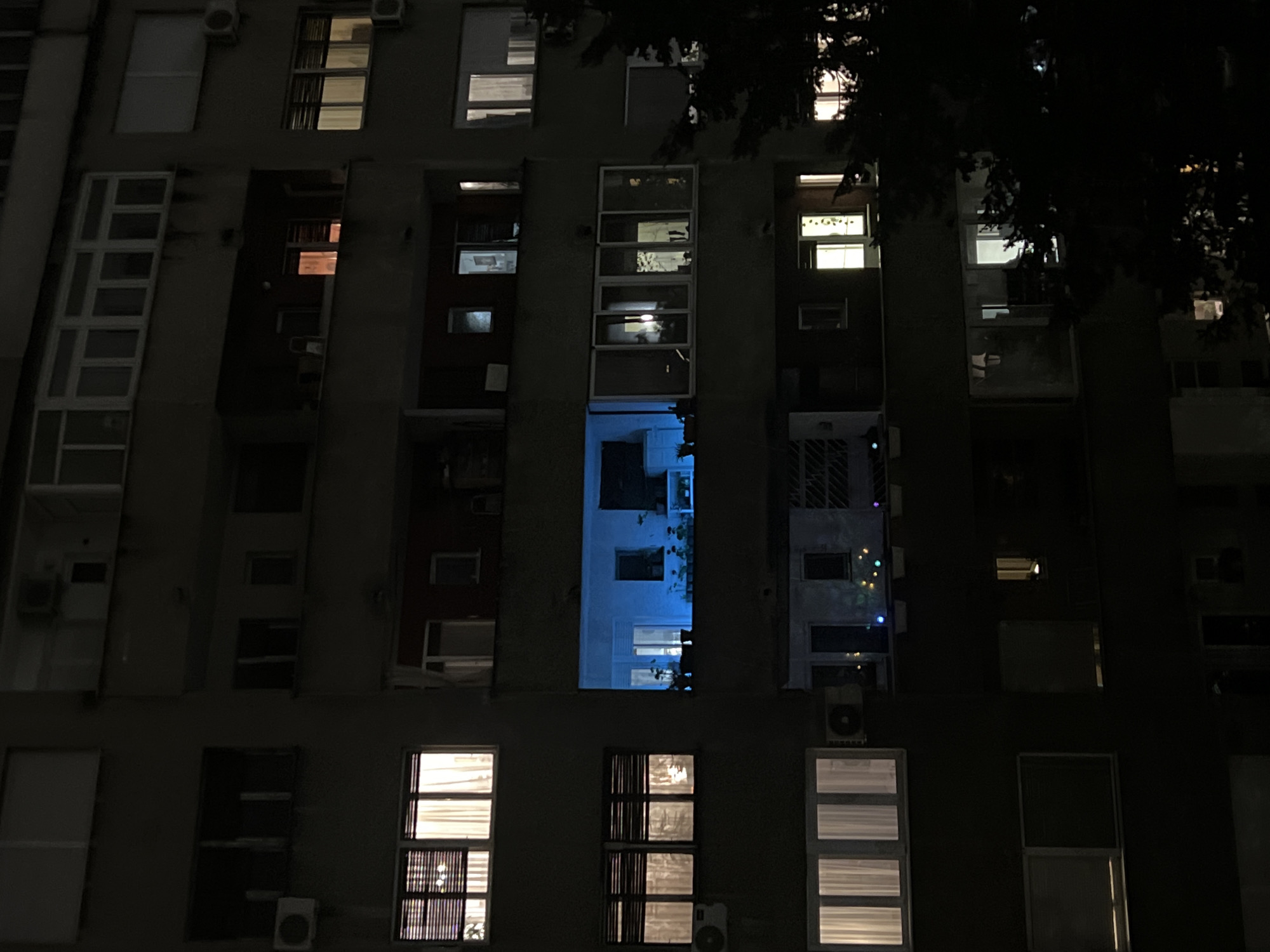 The image captures the facade of a multi-story apartment building at night in Belgrade, Serbia. The overall scene is dark, with various windows illuminated from within, suggesting occupied residences. The building features a uniform, somewhat modernist architectural style, with several rectangular windows of different sizes and configurations.
On the left side of the building, there are elongated, multi-pane windows that form enclosed balcony-like structures. Similar structures are visible in the central columns of the building. Below some windows, small outdoor air conditioning units are affixed to the wall.
A prominent feature is a large, rectangular window or open balcony in the center of the image, which emits a strong, distinct blue light, contrasting sharply with the warmer yellow and white lights coming from other apartments. Some windows show faint outlines of interior objects or curtains. On a balcony or enclosed area on the right side, colorful string lights are visible, adding a festive touch.
No people are discernible in the image, and there is no visible text. The atmosphere is quiet and still, characteristic of an urban residential area after dark. The varying patterns of light and shadow create a sense of individual lives within a communal structure.