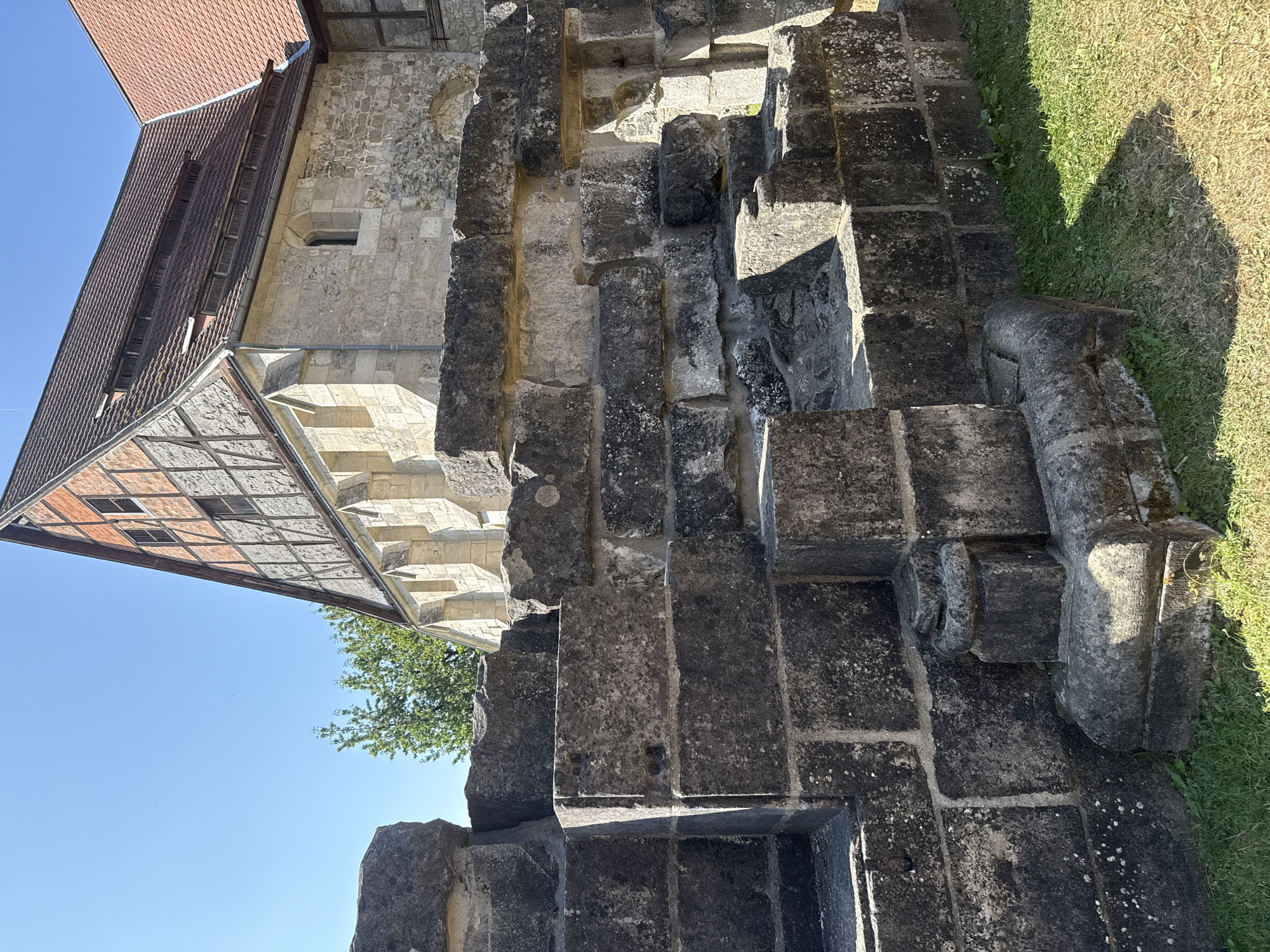 The image shows the exterior of a historic building, likely a church or monastery, in Walkenried, Germany. In the foreground, there is a weathered stone wall or ruin constructed from large, dark gray blocks. The blocks vary in size and are stacked unevenly, suggesting age and perhaps reconstruction. Some of the stones have visible lichen or moss growth. To the left of the stone structure, there is a patch of green grass.
Behind the stone structure, a portion of a building with a red-tiled roof is visible. The upper part of this building features a steeply pitched gable with exposed timber framing and brick infill, a style indicative of traditional German architecture. The lower part of the building is constructed from lighter colored stone blocks. The building has a large, prominent roof with dormer windows featuring louvers.
A clear blue sky dominates the background, with a few faint, dark specks that could be distant birds or dust motes. A green tree with lush foliage is partially visible behind the stone structure and to the left of the building. The sunlight suggests it is daytime, and the shadows indicate a sunny environment. There are no people visible in the image. The overall scene depicts a historical architectural site under a clear sky.