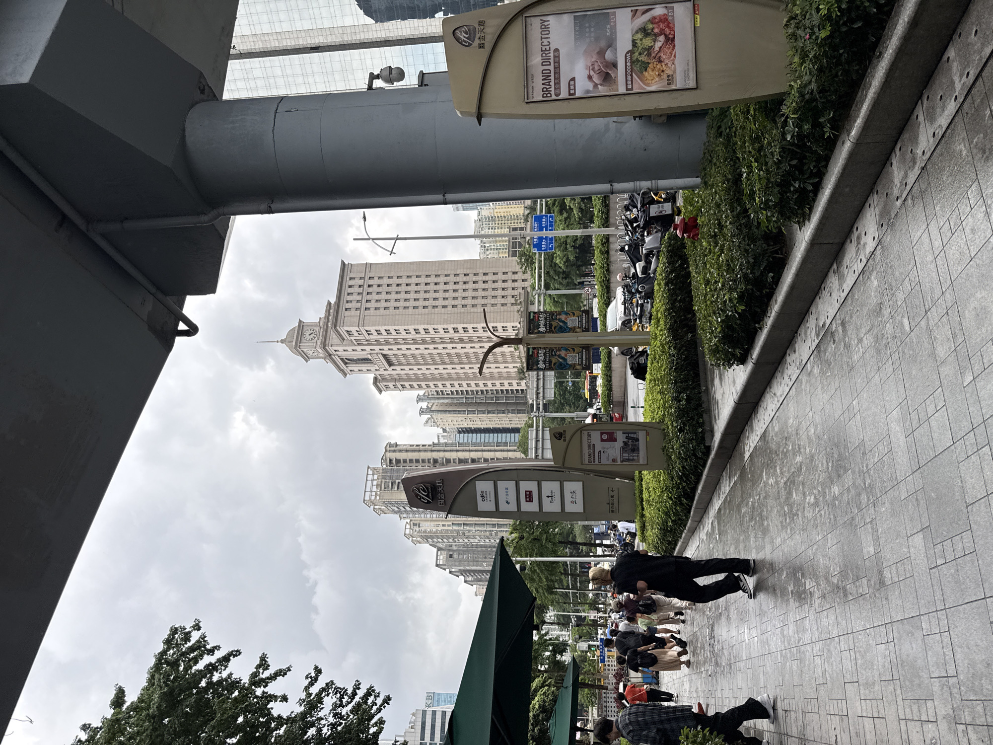 The image shows a sidewalk in Tianhe District, China, with a group of people walking. In the background, there are tall buildings, some with glass facades, and a clock tower. A large signpost with "ifc" and Chinese characters stands on the right side of the image, and a "BRAND DIRECTORY" is displayed on it. The sidewalk is paved with gray tiles and appears wet, suggesting recent rain. Green foliage is visible along the side of the sidewalk, and a red fire hydrant is positioned near a patch of bushes. The sky is overcast, contributing to a muted lighting condition. Several motorcycles and scooters are parked on the street beyond the sidewalk.
