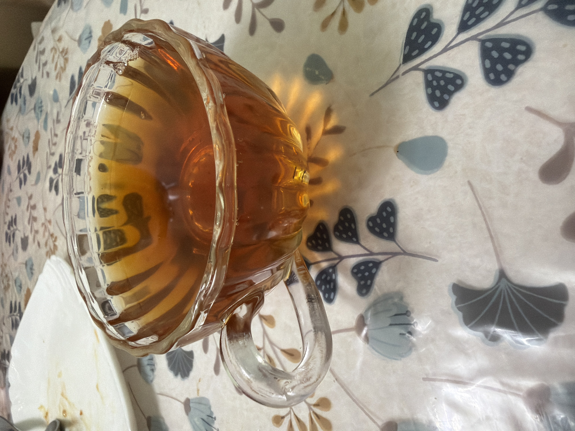 A clear glass teacup filled with amber-colored liquid sits on a table covered with a light-colored tablecloth featuring a floral and leaf pattern. The teacup has a ridged exterior and a clear glass handle. To the left of the teacup is a partially visible white plate with remnants of food. The background is softly blurred, but the tablecloth pattern is clearly discernible. The lighting suggests the scene is indoors.