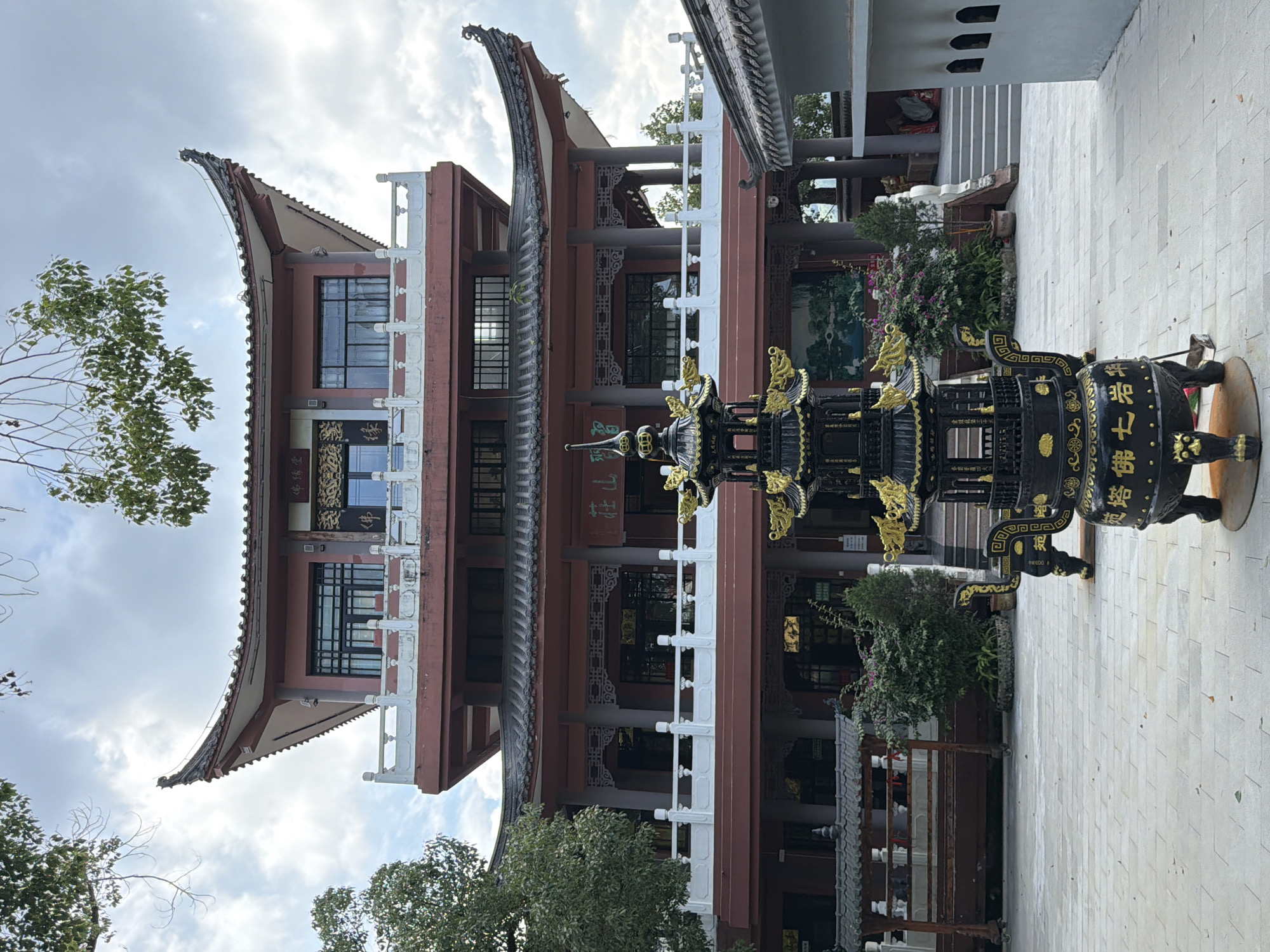 The image depicts an outdoor courtyard scene with a prominent multi-story, traditional Chinese-style building in the background. The building features multiple tiered roofs with upturned eaves, painted in red and grey tones, with white balustrades on its balconies. In the foreground, a large, ornate black incense burner with gold accents stands on the paved courtyard. The courtyard is paved with light-colored square tiles. To the left of the building, a covered structure with wooden benches is visible. Green trees and foliage surround the area. The sky is overcast with clouds. The scene appears to be a temple or cultural site. The inscription on the incense burner is in Chinese. Based on the building's architecture and the presence of traditional elements, this location is likely a temple or religious site within the Jiulongpo District of China.