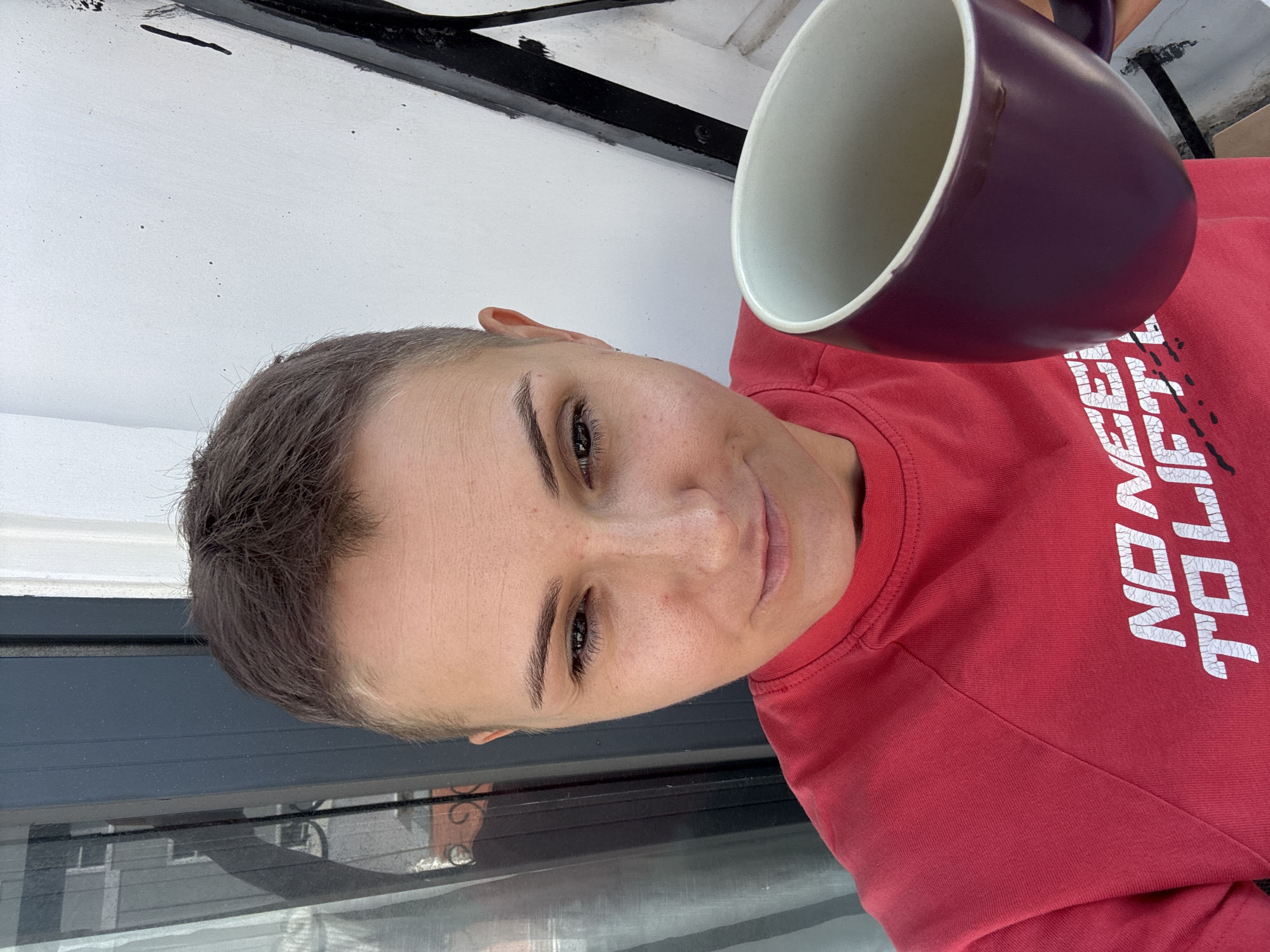 A close-up, selfie-style image shows a woman with short, dark hair holding a purple mug in her right hand, at chest level. She is wearing a red t-shirt with white text that appears to say "NO NEES TO LIFT". The woman's face is angled slightly down and to her left, with her eyes looking towards the camera. Her left eyebrow is slightly raised, and she has a subtle smile. The background is out of focus, but appears to be an outdoor setting with a grey wall and some white elements. There are no visible indications of the city of Nha Trang Ward, Vietnam.
