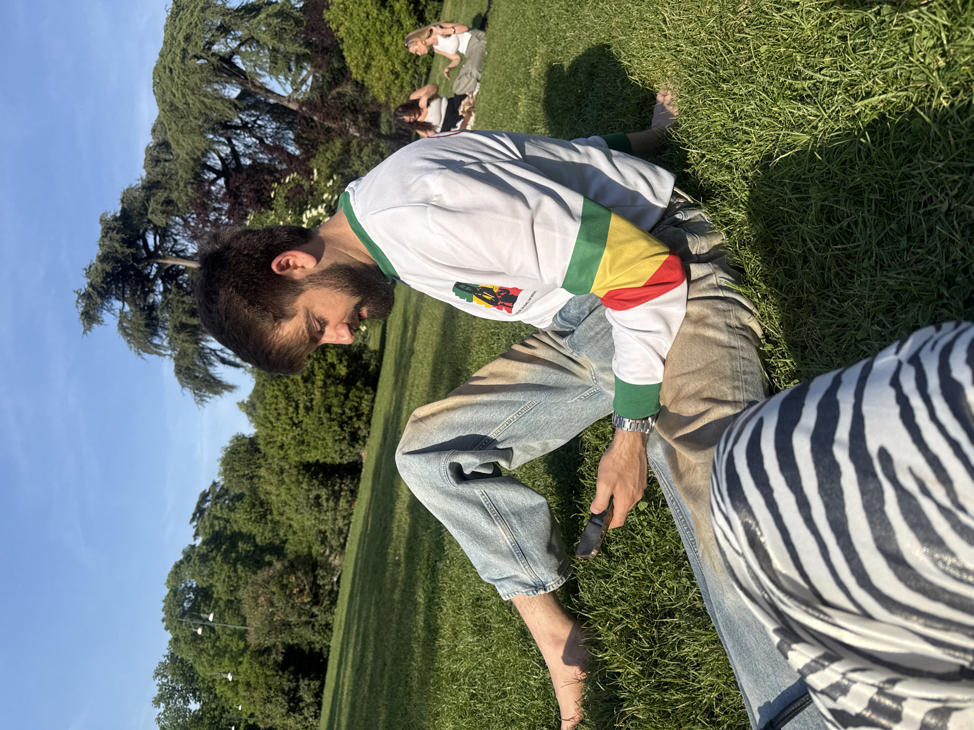 A sunlit outdoor scene captures people relaxing on a lush green lawn, likely in a park in Milan, Italy. The primary subject is a man with a beard and dark hair, seen in a squatting or kneeling posture on the grass, looking downwards. He is wearing a white long-sleeved shirt with green trim at the collar and cuffs, and a prominent graphic on the chest featuring red, yellow, and green stripes. The graphic includes text like "A Luta Continua," "BOB MARLEY," and "MARLEY 75," suggesting a connection to Bob Marley or reggae culture. He wears light-washed blue jeans and a silver-colored watch on his left wrist, and holds a dark smartphone in his right hand. His feet are bare on the grass.
In the mid-ground, two other blurry figures are visible on the grass, one appearing to be crouching or seated, and another further back, standing. The immediate foreground on the right shows a person's leg covered in a distinct black and white zebra-striped garment.
The setting is characterized by vibrant green grass and numerous large, mature trees, some coniferous and some deciduous, forming a dense backdrop under a clear, bright blue sky. Strong sunlight casts noticeable shadows, indicating a pleasant daytime, likely mid-morning or afternoon. The overall atmosphere appears relaxed and peaceful.