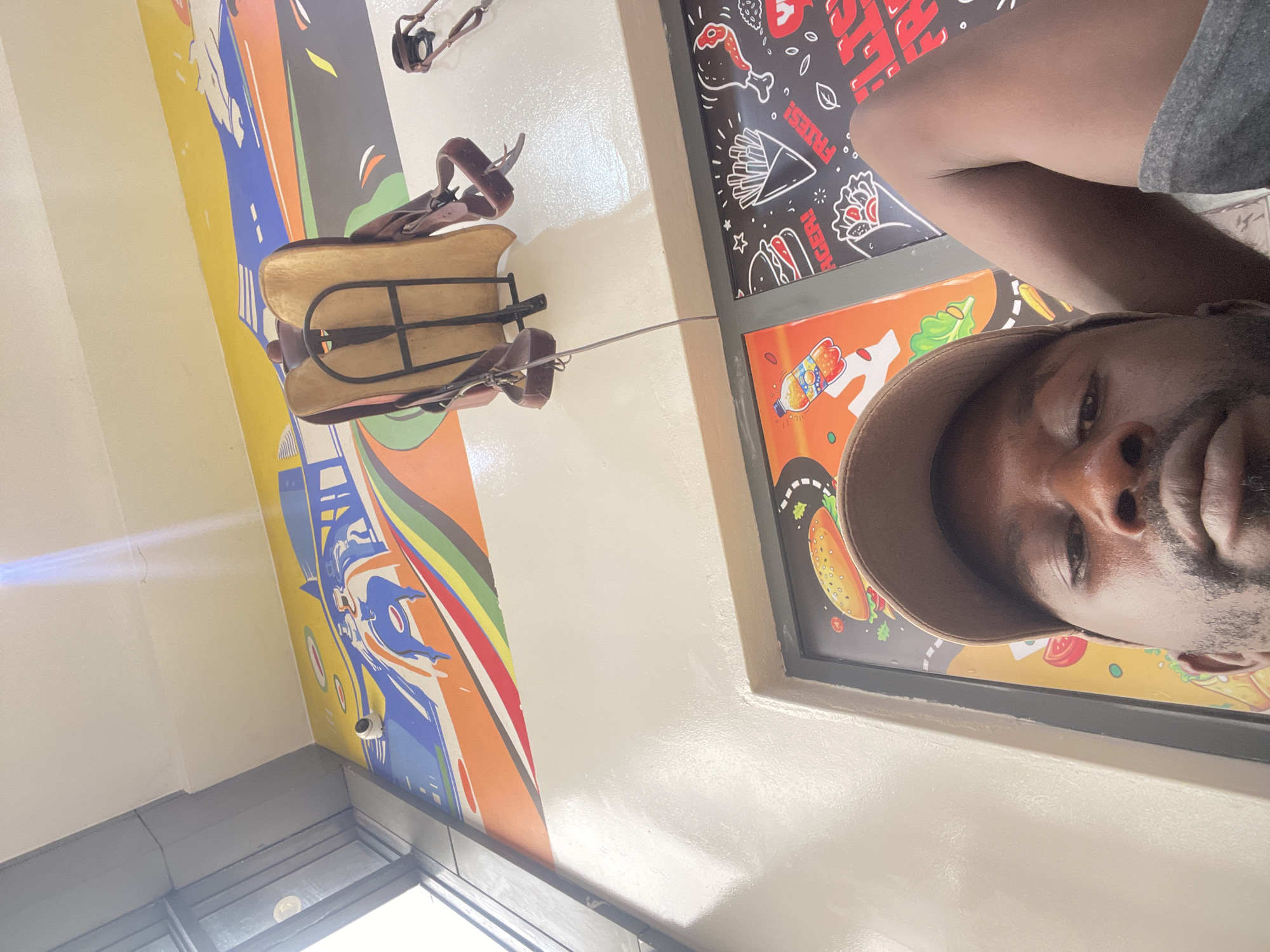 A man wearing a brown cap is taking a selfie. He is indoors, possibly in a restaurant or eatery, as there are fast food-themed decorations on the wall, including images of fries, burgers, and tacos. A child's horse saddle toy is also attached to the wall. The environment includes a window with daylight shining through. It is not possible to confirm the location of Rigachikun, Nigeria based on visual features present in this picture.