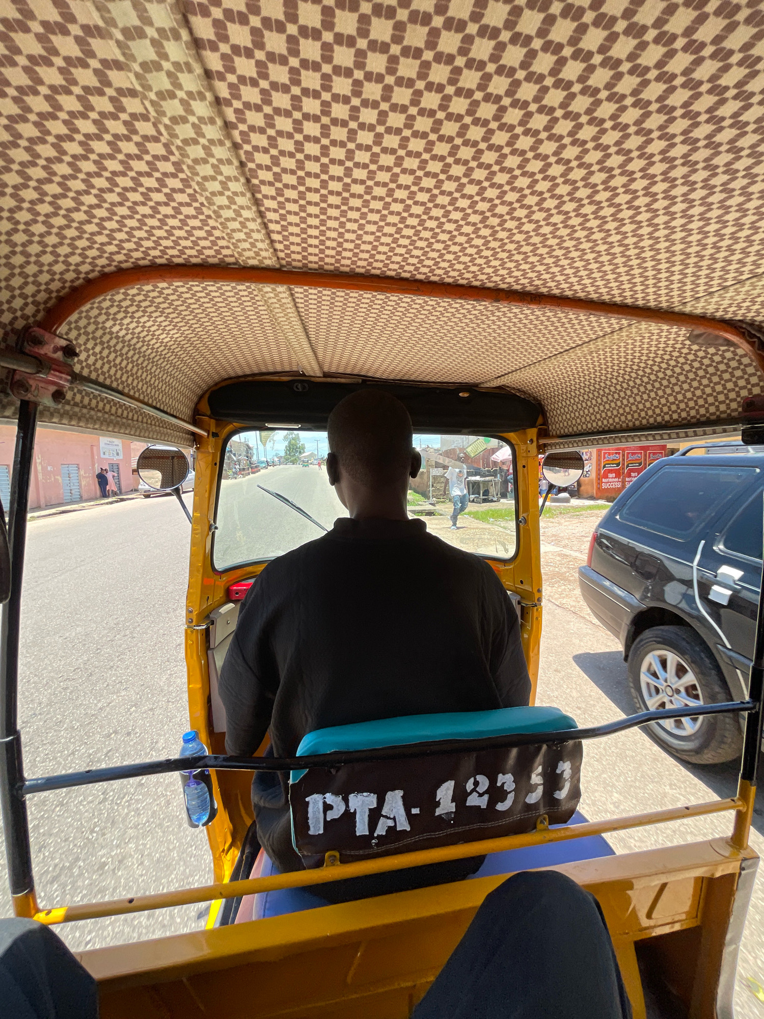 A rear-facing, eye-level shot captures the interior of a yellow auto-rickshaw as it drives through a street in Jos, Nigeria. The driver, a Black man wearing a black t-shirt, is seen from behind, operating the vehicle. A bottle of water is attached to a metal bar to the left of the driver. The roof of the auto-rickshaw is lined with a brown and beige checkered fabric. Through the windshield, a street scene unfolds with buildings, a black SUV, other vehicles, and pedestrians visible. A license plate on the back of the auto-rickshaw reads "PTA-12353". The overall impression is that of everyday transportation in a bustling city.