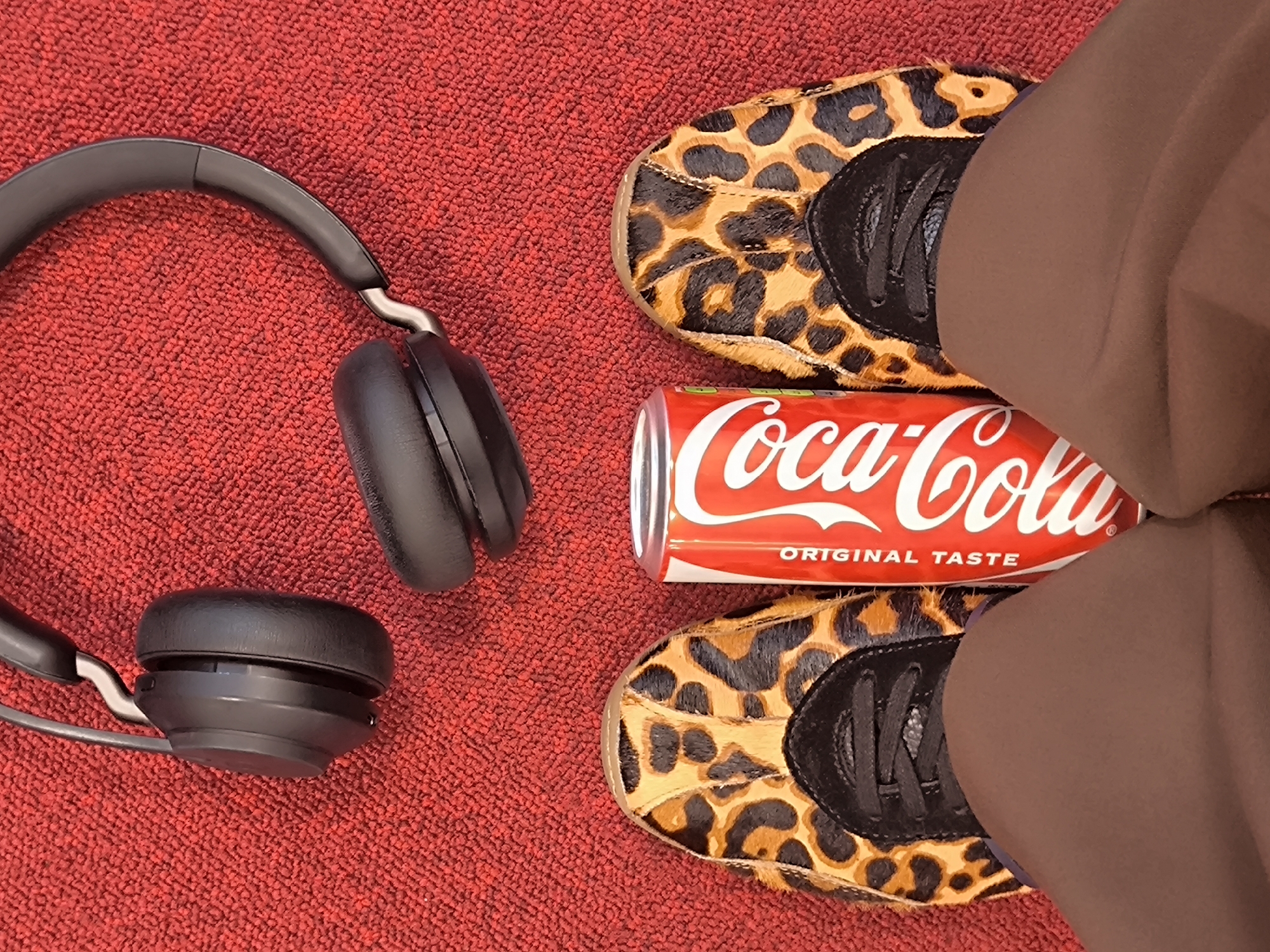 The image presents a still life composition on a red textured surface, possibly a carpet or rug. A pair of black, over-ear headphones is positioned on the left side of the frame. In the center, a Coca-Cola can is laid horizontally. To the right, there's a pair of shoes with a leopard print design, partially obscured by a brown-colored garment, likely pants. The location in the city of Riga, Latvia is not possible to infer from the image alone.