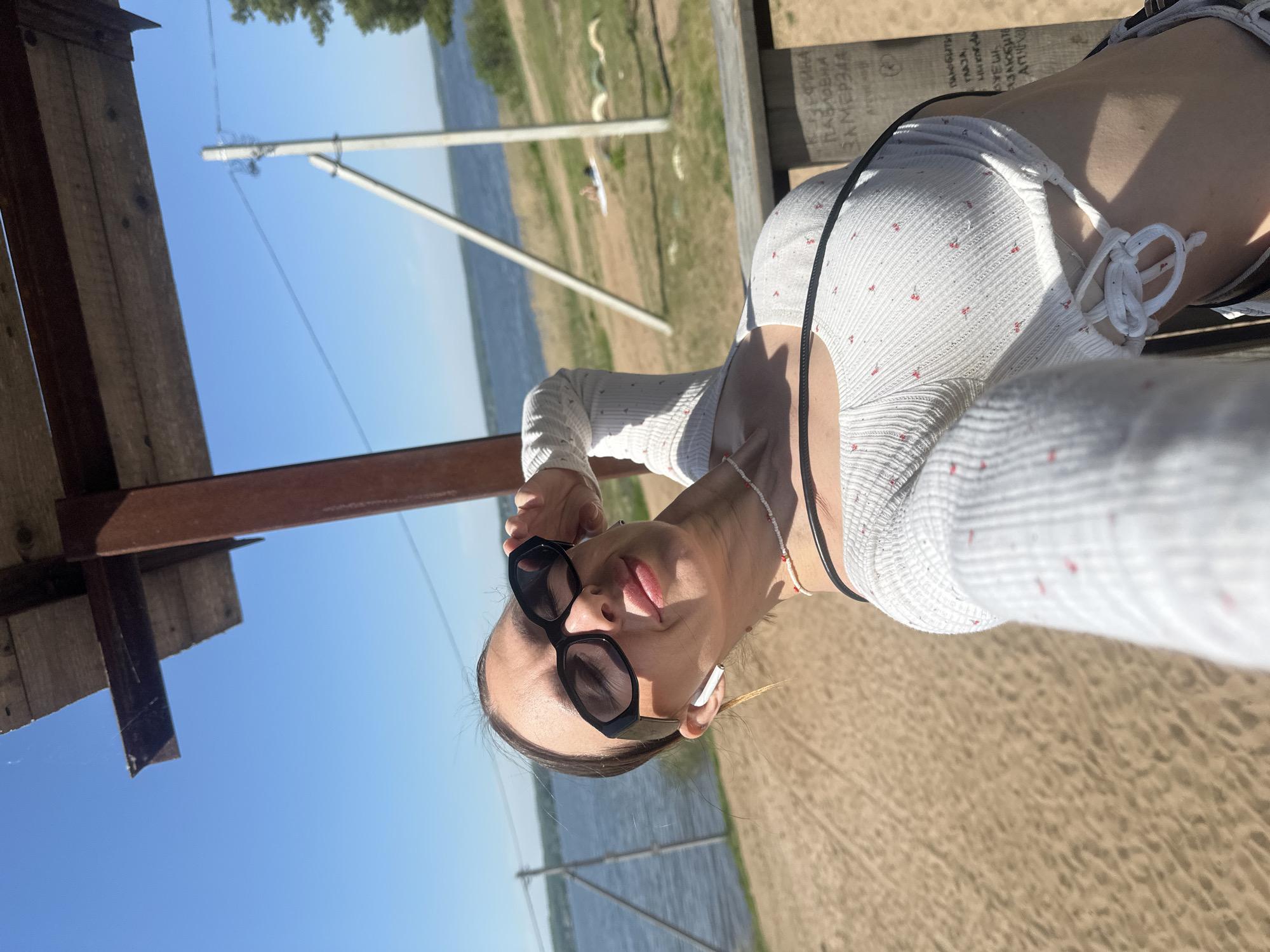 A woman is lying on a wooden surface in an outdoor setting, likely a beach or waterfront area, possibly near the city of Votkinsk, Russia. She is wearing sunglasses and a light-colored, patterned top. The backdrop includes water, a sandy area, and some vegetation. There is a wooden structure overhead, possibly a gazebo or a covered seating area.