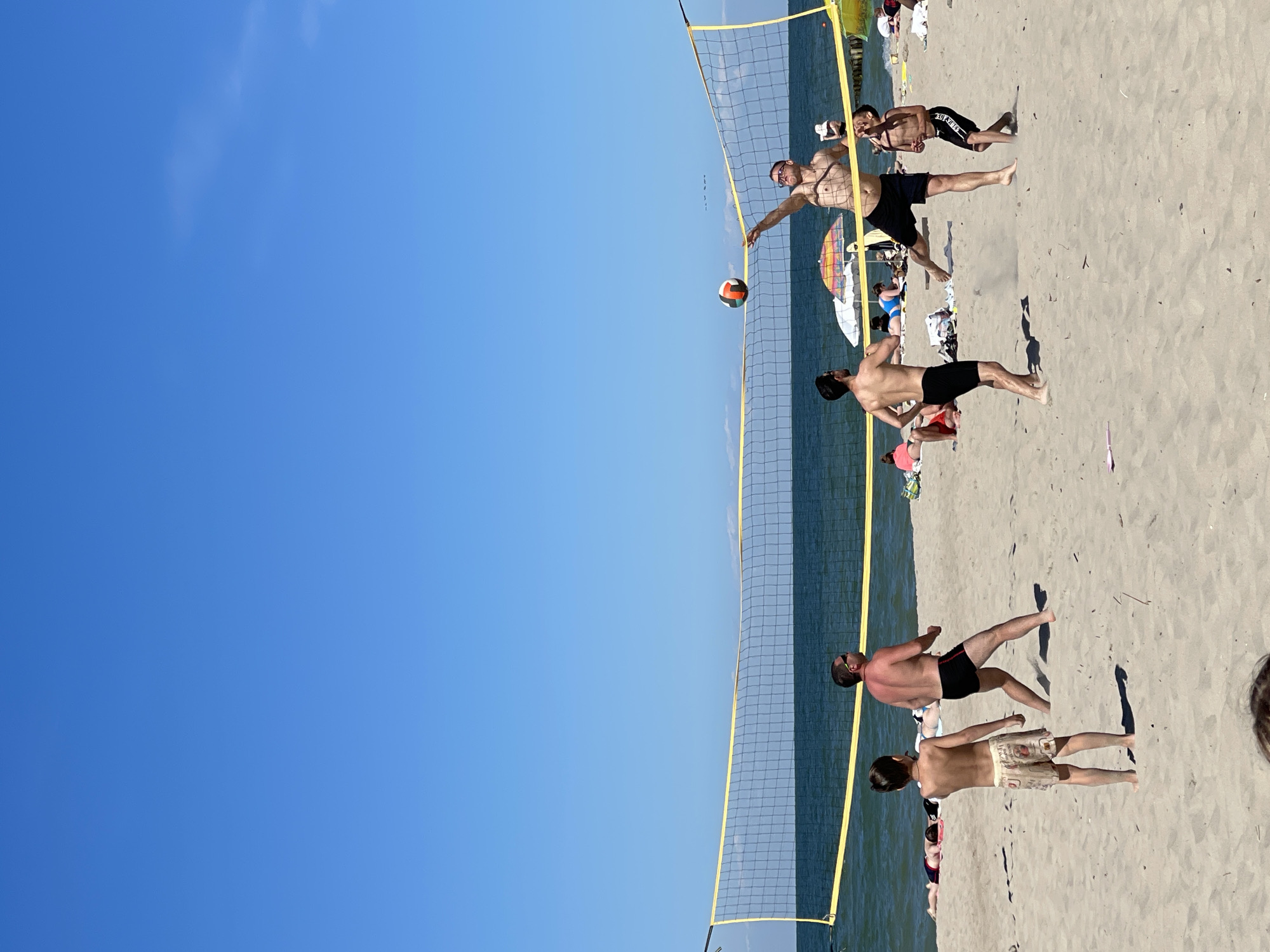 The image depicts a beach scene on a sunny day, with the Baltic Sea and a clear blue sky in the background. A group of people are playing beach volleyball. In the foreground, two young boys are standing on the sand. Further back, a group of men are actively engaged in the game. One man is jumping to hit the volleyball over the net, while others are positioned to receive or return the ball. Beach umbrellas and other beachgoers are visible in the middle ground, suggesting a public beach. The scene appears to be at a beach located in Zelenogradsk, Russia, a popular resort town on the Baltic Sea coast.