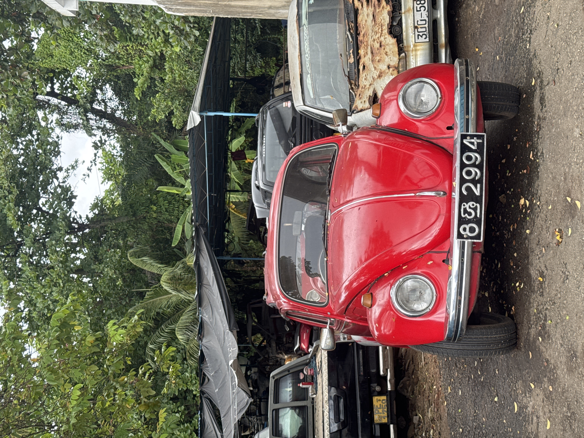The image shows a red Volkswagen Beetle parked on a road in Sri Jayewardenepura Kotte, Sri Lanka. The car is facing the viewer, and its license plate is visible. Other vehicles are parked behind the Beetle. The scene is partially shaded by a canopy structure, with trees and foliage visible in the background.