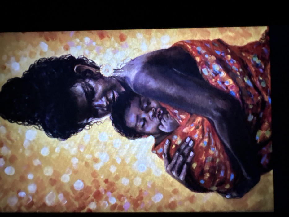 This image is a painting depicting a woman holding an infant.
**Subjects:**
* **Woman:** An adult female with dark skin tones, positioned facing slightly left, looking down towards the infant. Her dark, curly hair is styled in a high bun. She wears a prominent, decorative circular earring with a dangling element on her visible right ear. Her left shoulder and arm are visible, bare, and she uses both arms to cradle the baby.
* **Infant:** A baby with dark skin tones, held securely against the woman's chest and left shoulder. The infant's eyes are closed, indicating sleep or rest, and its right hand is near its chin.
**Objects:**
* **Fabric Wrap:** The infant is wrapped in a vibrant, multi-colored fabric, predominantly red-orange with scattered patterns of blue, yellow, and lighter orange shapes. This fabric also drapes around the lower part of the woman's visible arm and torso.
**Actions/Interactions:**
* The woman is holding the infant close to her body in a cradling manner, with her head inclined towards the baby.
**Environment:**
* **Background:** The background is abstract, featuring a warm yellow-orange hue with numerous soft, diffused circular or amorphous spots of white, yellow, and orange-red, suggesting a luminous or glittering effect.
**Location in the city of Jos, Nigeria:**
This image is an artistic rendering, specifically a painting, and does not depict a discernible physical location or real-world scene within the city of Jos, Nigeria. It is a studio or conceptual artwork.