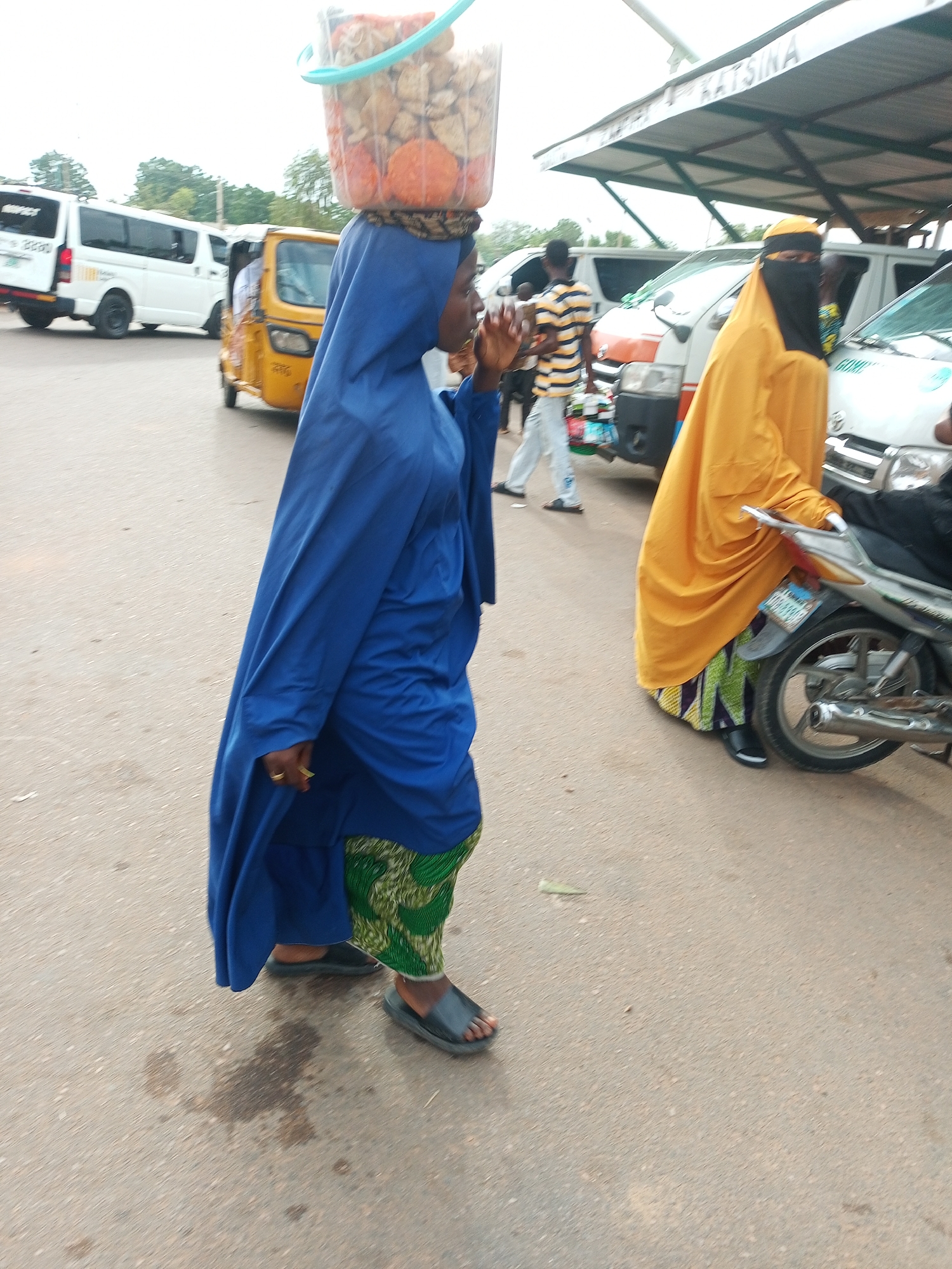 The image captures a street scene in Gombe, Nigeria, likely during daylight hours. The primary subject is a woman walking, carrying a large plastic bucket filled with food items on her head. She is wearing a blue abaya and a green patterned skirt, with black sandals on her feet. Her face is partially obscured by the blue fabric of her abaya. Beside her, another woman in a bright yellow hijab and black niqab walks towards a motorcycle.
In the background, several vehicles are visible, including a yellow tricycle and white minivans, suggesting a busy urban environment. One of the minivans appears to be a commercial bus. A covered awning with some lettering, partially visible as "KATSINA," is above the vehicles. The ground is paved asphalt, with some scattered debris. The overall impression is of a lively marketplace or transport hub, with individuals engaged in daily activities. There are no strong indicators of the weather, but the lighting suggests it is daytime. The activity depicted is simply people going about their business in a public space.