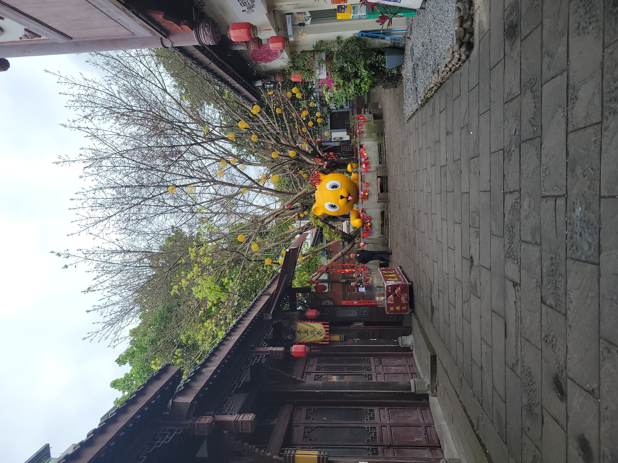 A vertical shot depicts a street scene in Hechuan, China. The foreground features a traditional dark wood building with intricate architectural details, including dark red doors and ornate carvings under the eaves. In front of the building is a vendor's cart and a person behind it. Above the cart is a tree, decorated with yellow lanterns and a cartoonish yellow bear with large eyes. The street is paved with large, rectangular stone slabs leading to the background, where there are other buildings, greenery, and decorations. The sky is overcast.