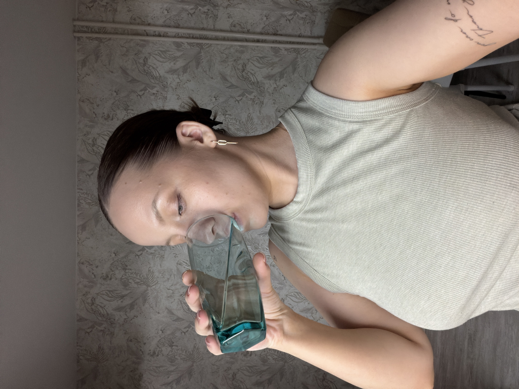 An indoor portrait features a person, likely a woman, drinking from a glass. Her dark hair is pulled back, and she wears a light-colored, ribbed short-sleeved t-shirt. A distinct gold-colored, rectangular bar earring adorns her visible ear. Her eyes are partially closed as she sips from a square-shaped glass with a teal tint, presumably containing water.
On her upper right arm, a tattoo is visible, consisting of partially legible text. Fragments like "man of" and "There was" can be discerned. The background wall is covered in a patterned wallpaper featuring a subtle floral or leaf design in muted gray and off-white tones. A plainer, darker gray wall section is visible to the left. The overall setting appears casual, with soft, ambient lighting. This scene takes place in Reshetnikovo, Russia.