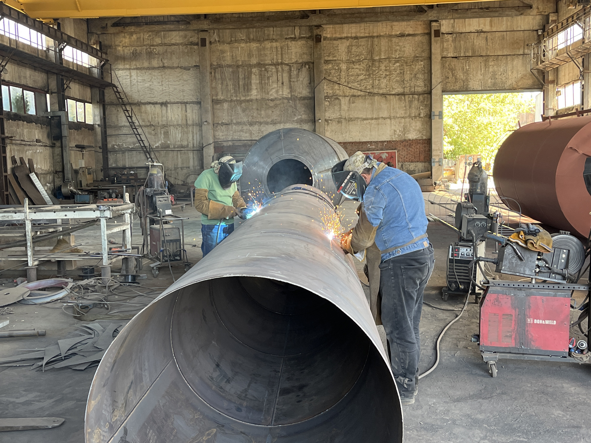 The image depicts a metal fabrication workshop, likely located in Taganrog City District, Russia, where two individuals are actively engaged in welding large metal pipes. The environment appears industrial, characterized by concrete walls and a high ceiling with a yellow overhead crane. In the foreground, a massive, unfinished steel pipe dominates the scene, with welding sparks emanating from its surface. Two welders, clad in protective gear including welding masks and gloves, are positioned at opposite ends of the pipe, contributing to its construction. One welder wears a green shirt and brown work gloves, while the other is dressed in a denim jacket and dark jeans. Various welding equipment, including welding machines and gas cylinders, are visible around the workshop, alongside stacks of metal sheets and other industrial materials. The background reveals additional large metal structures and a large opening that allows natural light to illuminate the workspace, with lush green foliage visible outside.