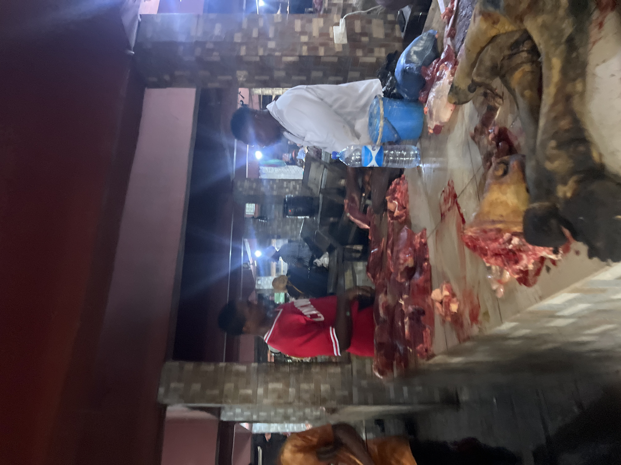 In Nkpor, Nigeria, within a bustling market environment, two men are observed behind a butcher's counter laden with raw meat. The man on the left, wearing a red shirt with white stripes, is actively engaged, while the man on the right, dressed in a white shirt, stands with his arms crossed. The counter displays a variety of meat cuts, some appearing to be whole carcasses or large portions, along with chopped pieces. A bottle of water and a blue bucket are also visible on the counter. The setting suggests a wholesale or retail butchery operation, with overhead lighting illuminating the scene and casting reflections. The overall atmosphere appears to be that of a working market, likely during operating hours.