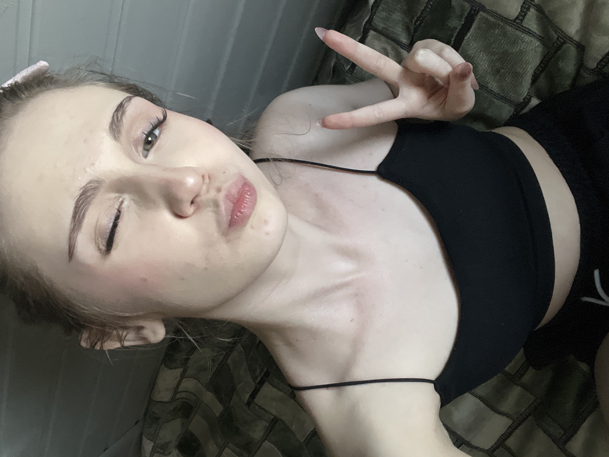 This media file shows a close-up, slightly tilted selfie of a young woman. She is lying down, with her head turned towards the camera. She has fair skin, light-colored eyes, and dark eyebrows. Her lips are slightly parted, and she is making a "peace" sign with her right hand, which is held up near her face. She is wearing a black tank top with thin straps and black shorts. The background appears to be a textured surface, possibly a bed or couch, with a patterned throw or blanket. The lighting suggests an indoor setting, possibly during the day given the brightness. Her expression is playful and relaxed. No text is visible in the image. The provided location context is Ashkadar, Russia.