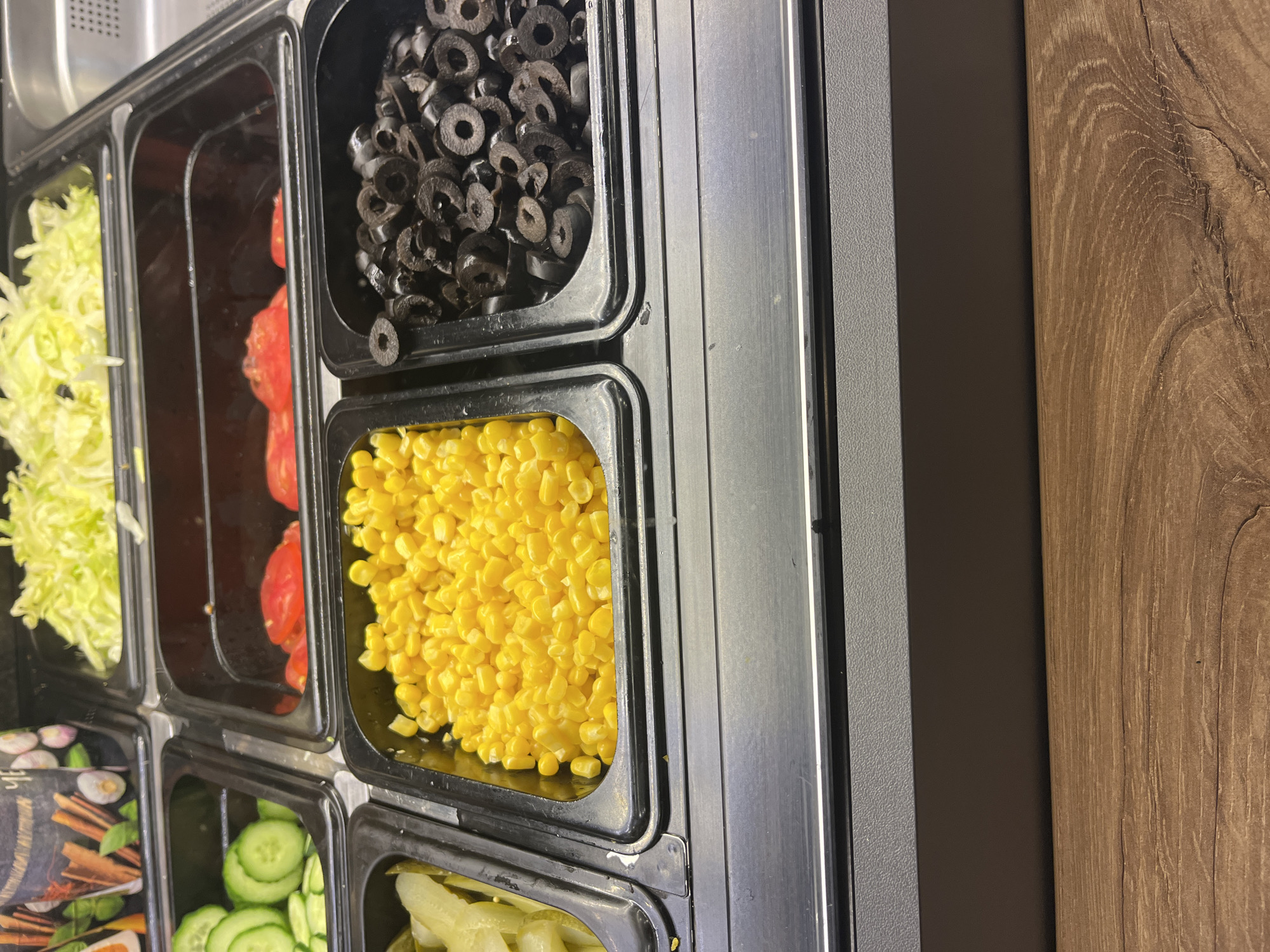 This image captures a close-up view of a food preparation line, likely in a sandwich shop, salad bar, or fast-food establishment. Various fresh ingredients are neatly displayed in rectangular containers made of black plastic or metal, housed within a shiny silver metal counter structure.
From left to right in the top row, the containers hold shredded light green lettuce, bright red sliced tomatoes (partially filled), and numerous sliced black olives. In the middle row, a container of vibrant yellow corn kernels is prominent. The bottom row displays sliced green cucumbers, and what appear to be pickled gherkins or possibly jalapenos. Another container in the bottom left corner features a decorative graphic with images of various spices and herbs, including cinnamon sticks, bay leaves, garlic cloves, and onions.
The silver metal framework extends to the right, adjacent to a dark brown vertical panel, which is next to a lighter brown wooden panel featuring a distinct wood grain pattern. On the far left, a textured gray surface is visible above the food containers.
The ingredients appear fresh and organized, suggesting a clean and well-maintained food service area. No people are visible, and no specific activity is taking place other than the presentation of the ingredients. There is no legible text visible on any of the containers or surfaces. Based on the provided context, the location is Burgas, Bulgaria, although the visual elements are characteristic of similar food service counters globally.