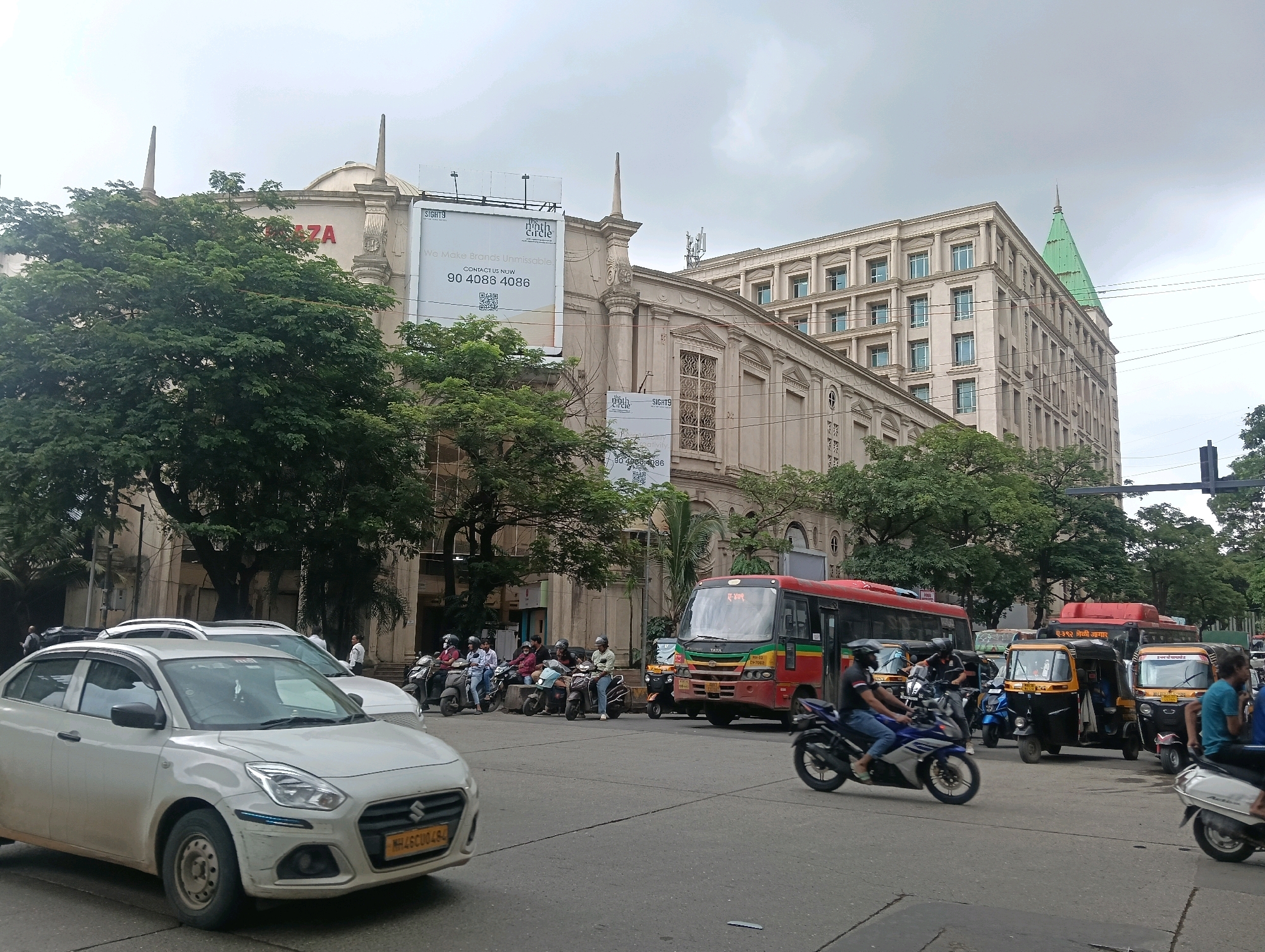 The image captures a street scene in Mumbai, India, likely a commercial or downtown area, with a focus on vehicles and buildings. In the foreground, a white sedan is parked. The middle ground features a red and yellow bus, several auto-rickshaws, and multiple motorcycles with riders. A group of people are gathered near the motorcycles. In the background, a grand, multi-story building with classical architectural elements and a green spire dominates the skyline. Another building with large windows and columns is also visible. A billboard advertising "SIGHTS" and "the ninth circle" with a phone number and QR code is mounted on one of the buildings. The sky is overcast, suggesting a cloudy day. The street appears to be a busy intersection or thoroughfare.