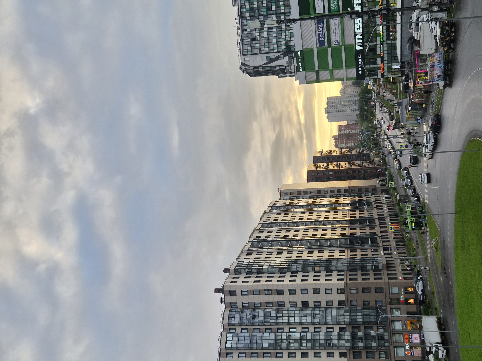 The image depicts a street scene in Kudrovo, Russia, with a focus on modern residential buildings and commercial establishments. The perspective is from an elevated viewpoint, looking down onto a busy street.
In the foreground, a well-maintained green grassy area is visible. The street itself is paved and features multiple lanes of traffic. Cars, including a white truck, are driving and parked along the road. Pedestrians are seen walking on sidewalks and crossing the street.
Dominating the scene are large, multi-story residential buildings with numerous windows. These buildings exhibit contemporary architectural styles. Adjacent to the residential structures, commercial buildings with prominent signage are present. Some of these signs indicate businesses such as "Fitness Life" and "Pedant".
The sky above is overcast with clouds, but shafts of golden sunlight break through, indicating either sunrise or sunset. The overall atmosphere of the scene is urban and active, with a blend of residential and commercial activity. The scene appears to be captured in the daytime, given the lighting conditions.