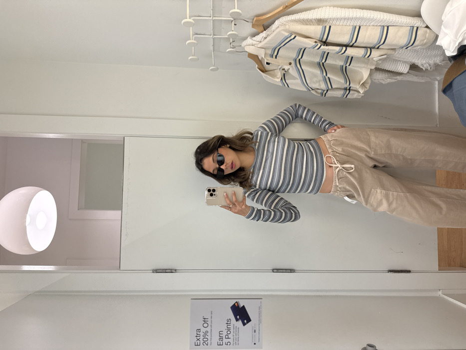 In a dressing room, a woman poses for a mirror selfie. She is wearing sunglasses, a striped long-sleeved shirt, and beige pants with a drawstring waist. A light fixture illuminates the left side of the frame, while clothing hangs on a rack above her. A sale poster is affixed to the door. The overall scene is well-lit and clean.