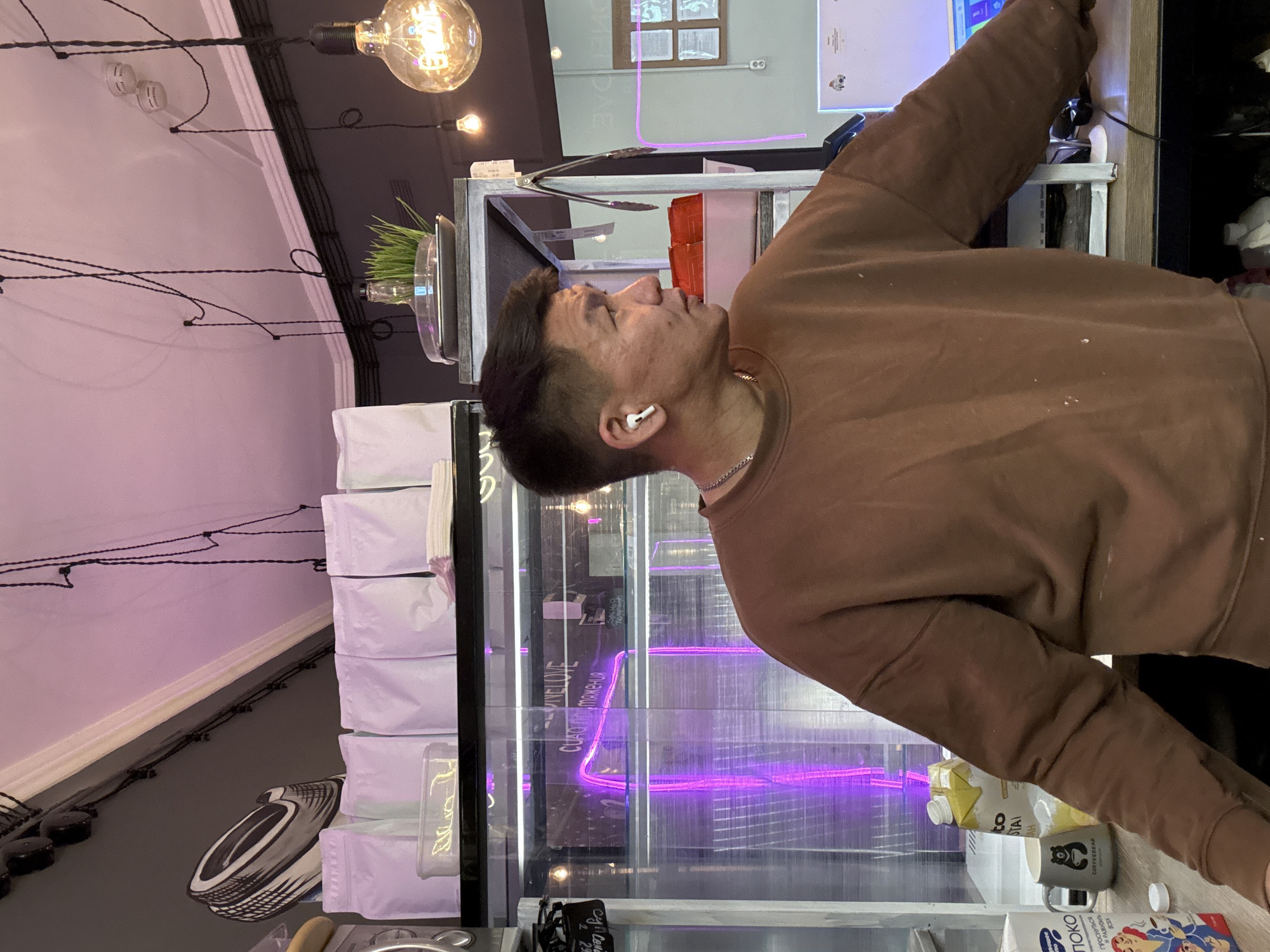 The image shows a man in a brown sweatshirt looking into a glass display case. He is wearing an earbud and a necklace. The display case appears to be in a retail or café setting. The case contains various products including some in white bags, a neon purple sign with Russian text (reading "PIXELOVE CUAT подтяжения"). There is also a container of wheatgrass visible on top of the case. On the counter in front of the case are a carton of soy milk, a mug, and another carton with a cartoon design. The lighting in the scene is a mix of natural and artificial light with an exposed filament bulb hanging from the ceiling. The ceiling is partly purple, partly dark gray, and partly light gray.
Based on the Russian text and the style of the scene, it is highly probable that this is taken in Moscow, Russia.
