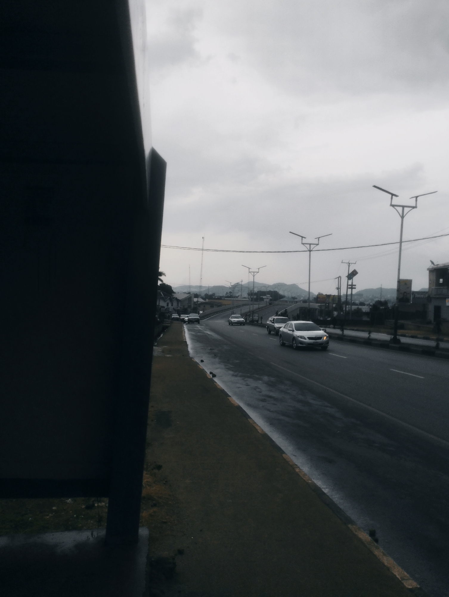 The image depicts a road in the city of Jos, Nigeria, under an overcast sky. The road is wet, indicating recent rain, with visible reflections on the asphalt. Several cars are driving on the road, including a silver sedan in the foreground. On the left side of the road, there is a grassy verge and a large, dark structure that appears to be part of a building or a shelter. In the background, there are buildings, streetlights, and hills. The overall scene suggests a typical day in a Nigerian city, with traffic moving along a main thoroughfare.