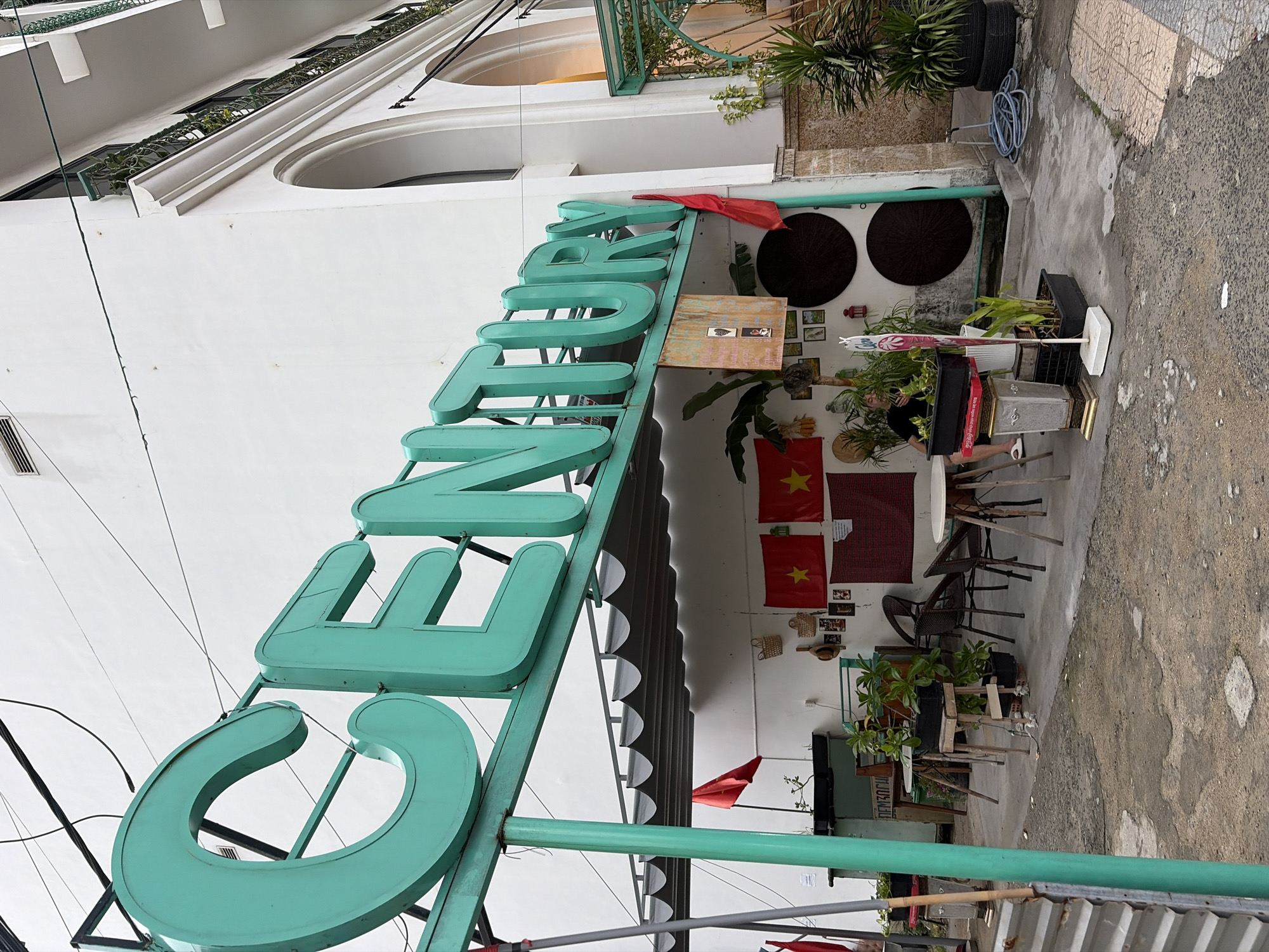 This image shows a street-level view of a building with a prominent teal-colored "CENTURY" sign. The building's facade is white, with arched windows and balconies visible on the upper floors. A covered outdoor seating area is situated in front of the building, featuring tables and chairs. Vietnamese flags are displayed on the wall behind the seating area, along with framed pictures and decorative items. Greenery, including potted plants and a palm tree, is scattered throughout the scene. A person wearing a black shirt and shorts is seated at one of the tables. The ground in front of the building is paved with concrete and asphalt. The overall impression is that of a cafe or restaurant with a distinct visual identity. The sign and flags suggest the location is likely in Vietnam, and given the context provided, it is identified as being in the city of Đà Nẵng.