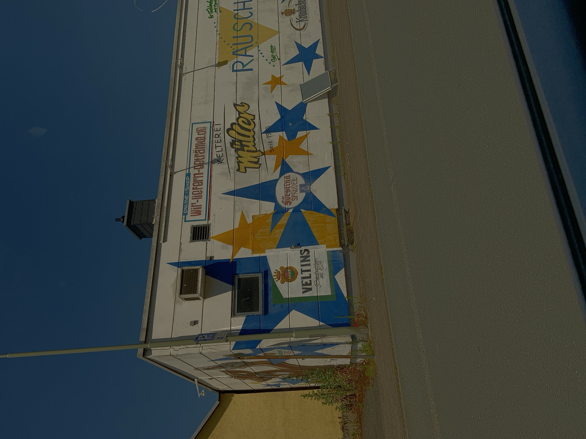 The media file shows a mural painted on the side of a building in Nauborn, Germany. The mural features a variety of text and graphics, including stars, beer logos, and slogans. The dominant text elements include "RAUSCH," "Miller," "Forstina SPRUDEL," and "VELTINS." There is also a website address, "Wir-liefern-getränke.de," and the phrase "ONLINE SHOP." The building appears to be a commercial establishment, possibly a beverage distributor given the prominent beer logos. The mural is colorful, with blues, yellows, and reds used prominently. The sky in the background is a clear, deep blue, suggesting a sunny day. The building itself is white, with some architectural features like an air conditioning unit and a window visible. The image is taken from a slightly low angle, looking up at the building. There is also a paved area and some greenery visible along the side of the building, indicating an outdoor setting. The overall impression is one of a brightly decorated commercial space under a clear sky.