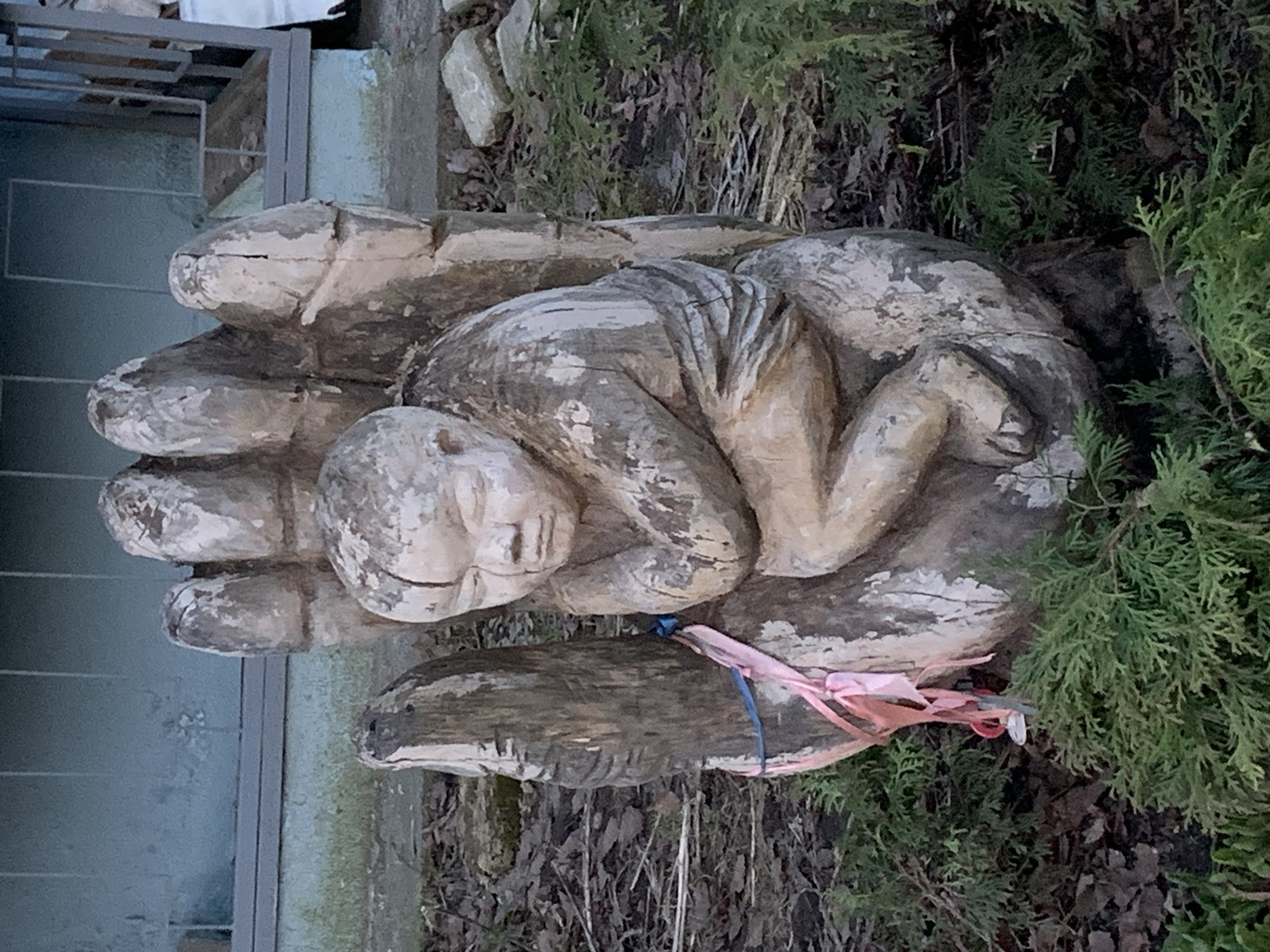 A weathered wooden sculpture depicts a small, human-like figure curled in a fetal position within the palm of a larger, stylized hand. The sculpture is made of light-colored wood, possibly oak or pine, and shows signs of aging with visible cracks, distressed texture, and patches where the original lighter color contrasts with darker, weathered areas. The figure's face is serene, with closed eyes, conveying a sense of peace or deep sleep.
The sculpture is situated outdoors, likely in a garden or yard. To the right, there are lush green coniferous shrubs or trees, while the ground around the base is covered with dry, fallen leaves and sparse brown vegetation, suggesting late autumn, winter, or early spring. A pink ribbon and a thin blue string are tied around the lower part of the hand, near the wrist, adding a touch of color to the otherwise natural and muted palette. In the upper left background, part of a building or structure with a glass panel and metal framing is visible, hinting at a residential or landscaped setting. The lighting appears soft and diffused, indicating either an overcast day or indirect sunlight. There is no visible text in the image. The sculpture evokes themes of protection, vulnerability, and care. This scene is located in Chaikovsky, Russia.