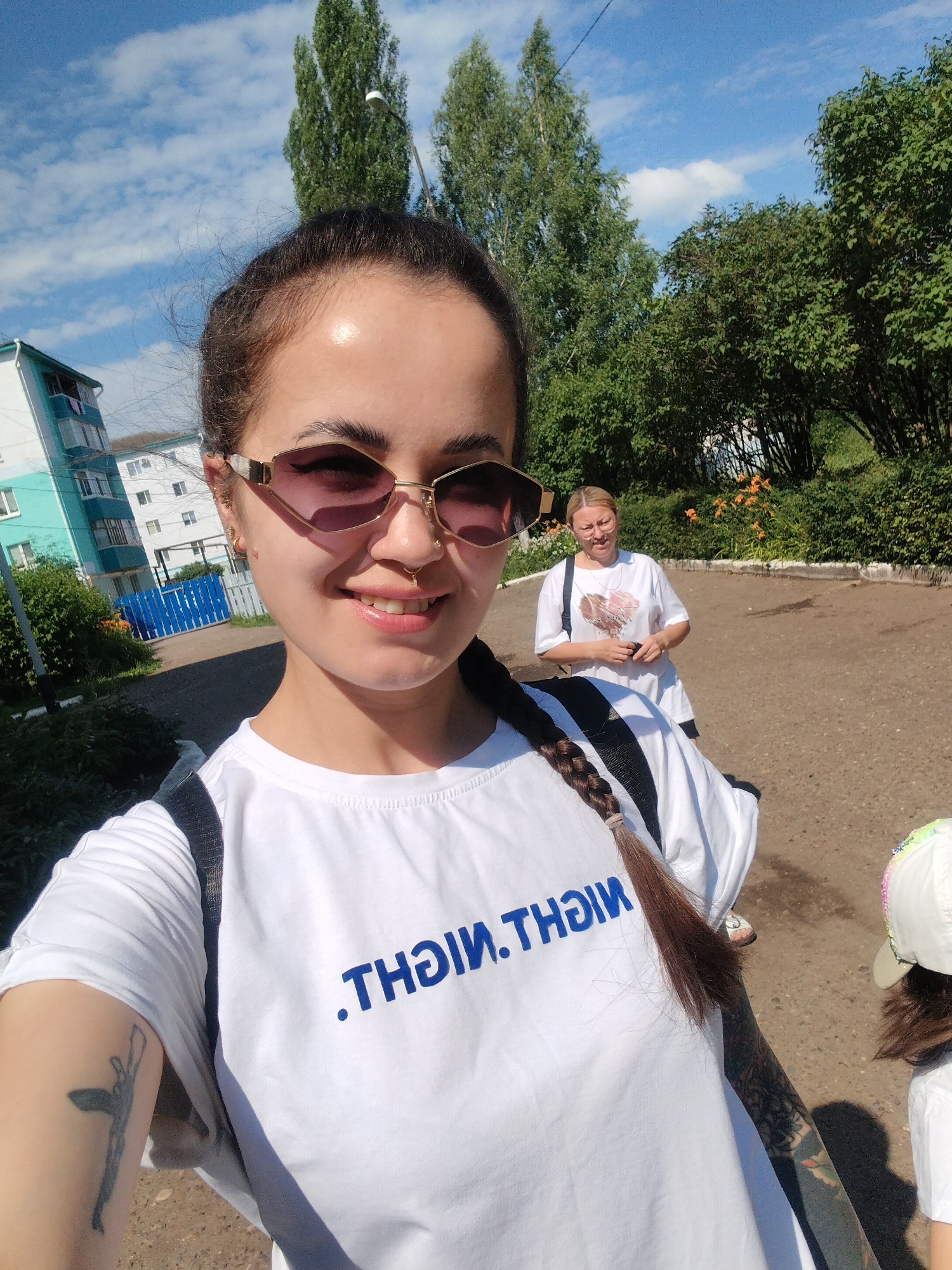 In Yanaul, Russia, on a sunny day, a young woman with dark hair pulled back, wearing large sunglasses and a white t-shirt, is taking a selfie outdoors. Her t-shirt has blue text that appears to be reversed. A woman in the background, also wearing a white t-shirt and glasses, holds a phone. A child in a white cap is partially visible to the right. The environment includes trees, a blue fence, a building, and a paved walkway. The woman has tattoos on her arm.