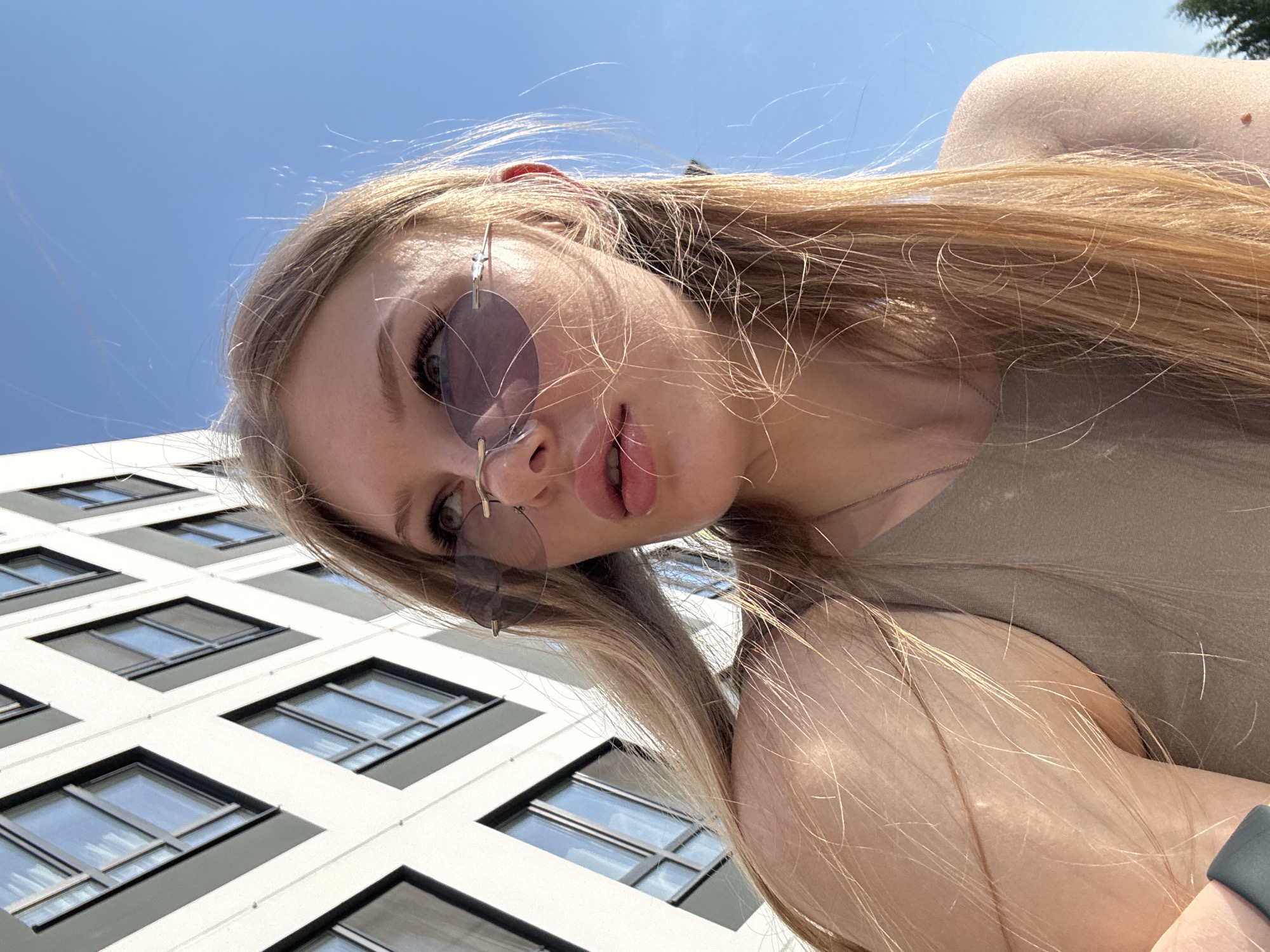 A low-angle, close-up selfie of a young, fair-skinned woman with long, blonde hair wearing round, tinted sunglasses. She is looking directly into the camera with a slight downward tilt of her head. Her lips are slightly parted.
Behind her, the lower portion of a modern building with multiple rows of windows is visible. The building is light in color with dark window frames. The sky above the building is clear and blue. This photograph was taken in an outdoor setting within Perm, Russia, since it was stated as the location.