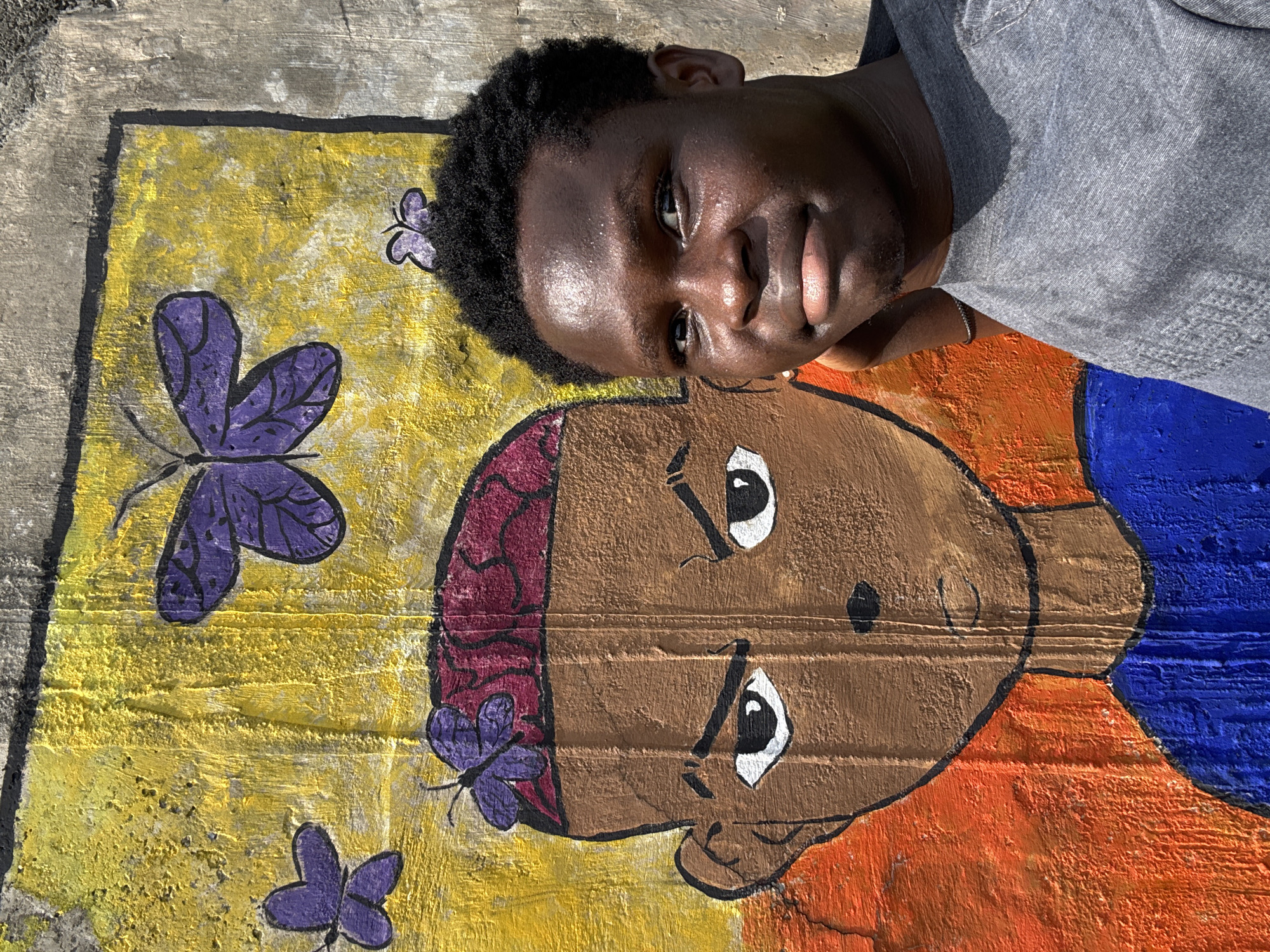 A young Black man poses lying down next to a vibrant mural painted on a concrete surface. He looks directly at the camera with a gentle smile, his face illuminated by bright sunlight which casts strong highlights on his skin. He has short, dark, curly hair and wears a grey t-shirt, also partially lit by the sun.
The mural, visible on the ground beside him, features a stylized human head and upper body. The painted figure has brown skin, large expressive white eyes with black pupils, and a small mouth. A reddish-purple wavy pattern is depicted on the side of its head. The figure's clothing or lower body is rendered in distinct sections of vibrant orange and blue. The background of the mural around the head is painted yellow and is adorned with several purple butterflies, outlined in black with visible veins on their wings. One large butterfly is positioned towards the upper left, while smaller ones are scattered, including one near the painted figure's ear. The concrete surface beneath the paint shows texture and some wear.
No text is visible in the image. The bright lighting suggests a sunny outdoor setting, consistent with the provided location context of Ihiagwa, Nigeria. The man appears relaxed and pleasant.