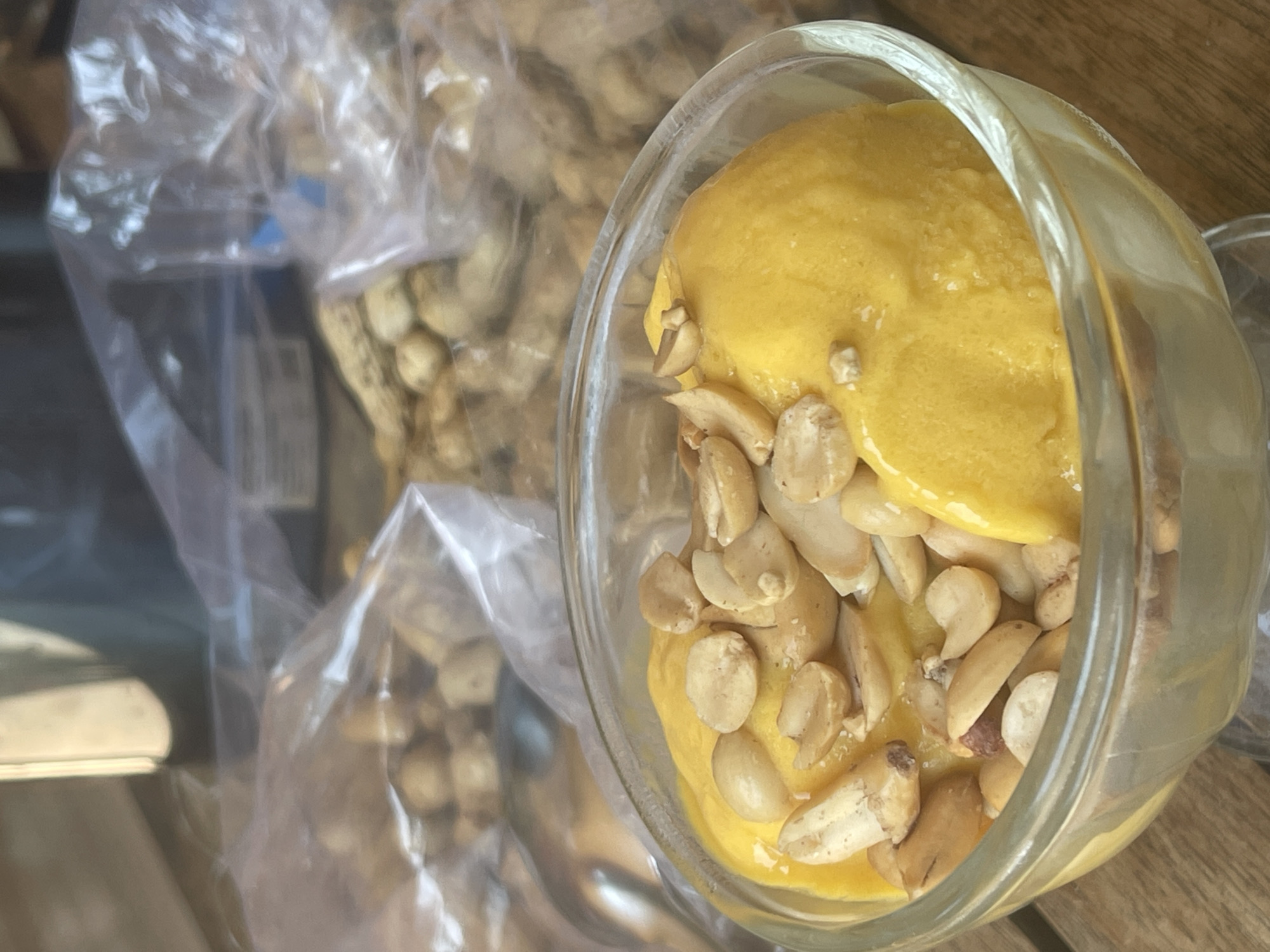 A close-up shot shows a clear glass bowl filled with bright yellow ice cream, possibly mango flavored, topped with a generous portion of roasted peanuts. The bowl rests on a wooden surface. In the blurred background, two clear plastic bags filled with more peanuts are visible. The overall scene suggests a casual outdoor setting, likely a meal or snack. The image does not contain information about the specific location in Phan Thiet Ward, Vietnam.