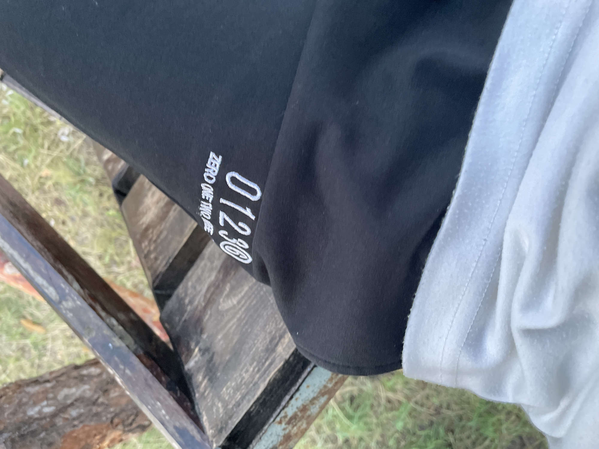 The image shows a close-up of clothing, likely a black garment with white lettering and a white garment. The black garment has "01230" and "ZERO ONE TWO" printed on it in white, stylized text. The white garment appears to be a t-shirt or similar top, folded over the black garment at the top right of the image. The clothing is resting on a wooden surface, possibly a bench or pallet, which is weathered and appears to be outdoors. The background is blurred but suggests an outdoor setting with grass and some natural elements. The lighting is consistent with daytime. No people are visible, and the focus is entirely on the textiles and the wooden surface.