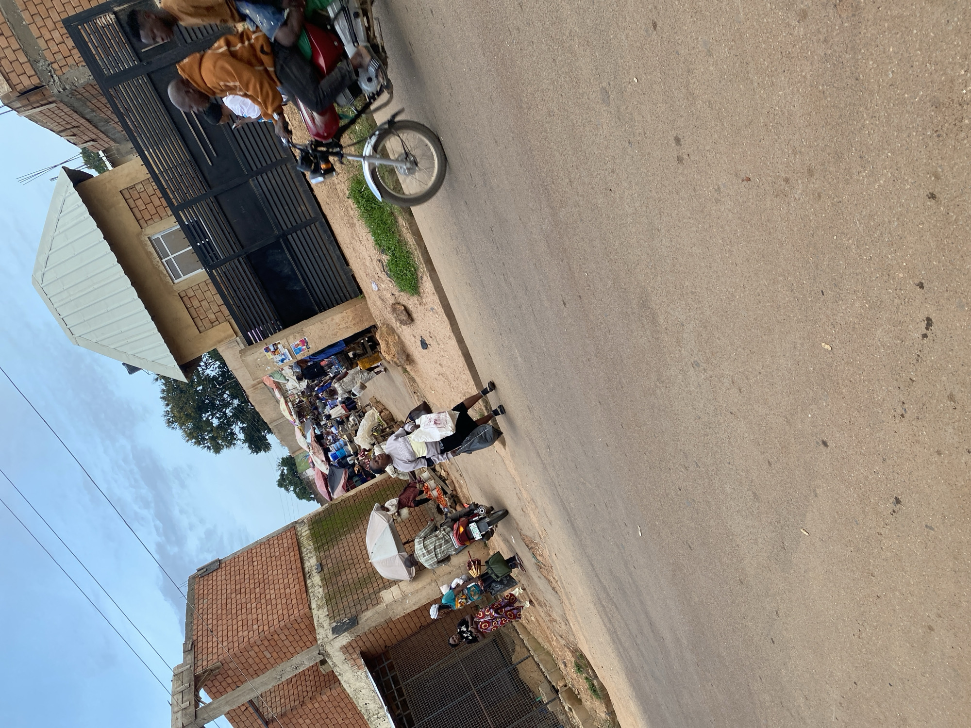 The image depicts a street scene in Bukuru, Nigeria, with a focus on a busy market area and an adjacent street. In the foreground, the asphalt road is visible, with a motorcycle in motion on the right side. The motorcycle is red and green, and two men are riding it, the driver wearing an orange hoodie and the passenger an orange shirt. The background reveals a bustling market with numerous stalls, umbrellas, and people. Several individuals are walking and interacting within the market. On the left side of the image, an unfinished brick building stands next to the market. The sky is partly cloudy.