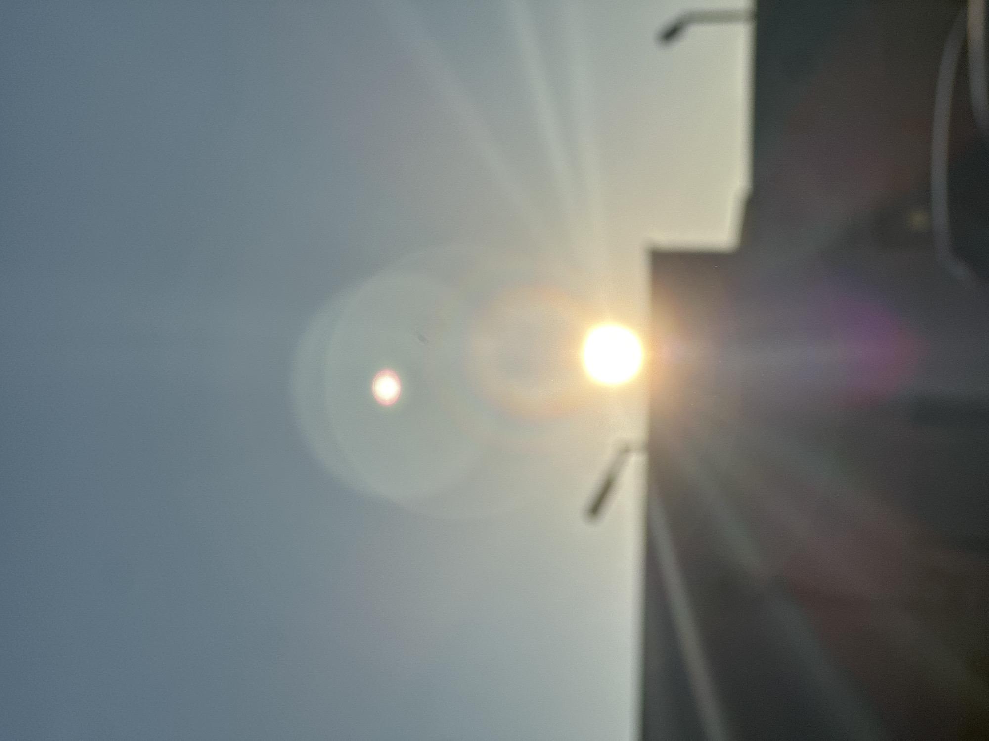 The image shows a bright sun shining through a building in Sofia, Bulgaria. The building is on the right side of the frame and obscures part of the sun. Lens flare is visible in the image, creating a circular artifact to the left of the sun. The sky is a pale blue color. The image is slightly out of focus.