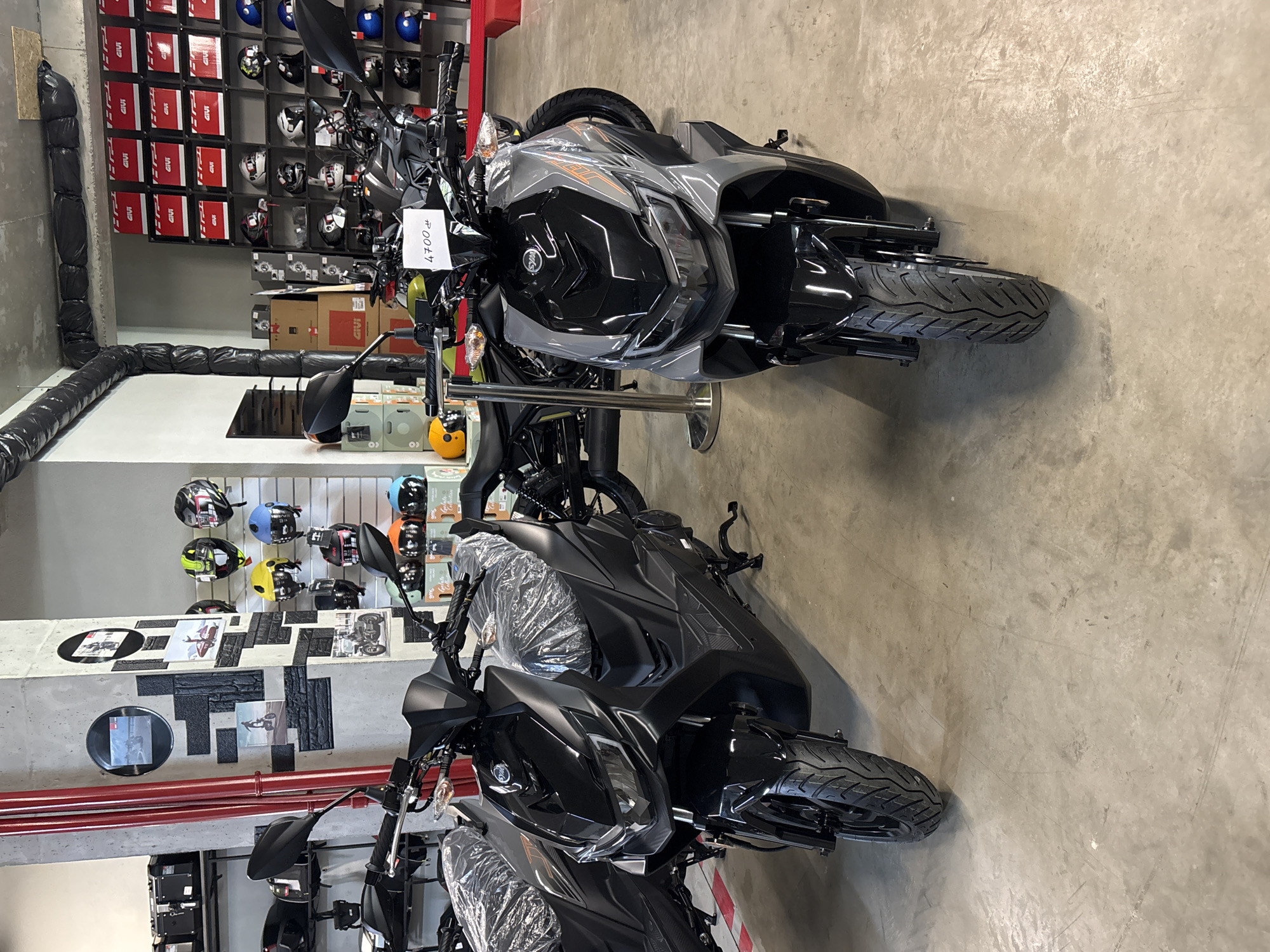 This is a photograph of a motorcycle showroom in Tbilisi, Georgia. Two scooters are prominently displayed in the foreground. The scooter on the left is black and covered in protective plastic, while the scooter on the right is gray and black, also with some plastic wrapping. A price tag reading "4700 ₾" is attached to the handlebars of the gray scooter.
In the background, shelves are stocked with various motorcycle helmets in different colors and styles. Some boxes, possibly for motorcycle accessories, are also visible. A red pipe runs vertically along a concrete pillar on the left side of the image. The floor appears to be concrete. The overall environment suggests a retail space for motorcycles and related gear.