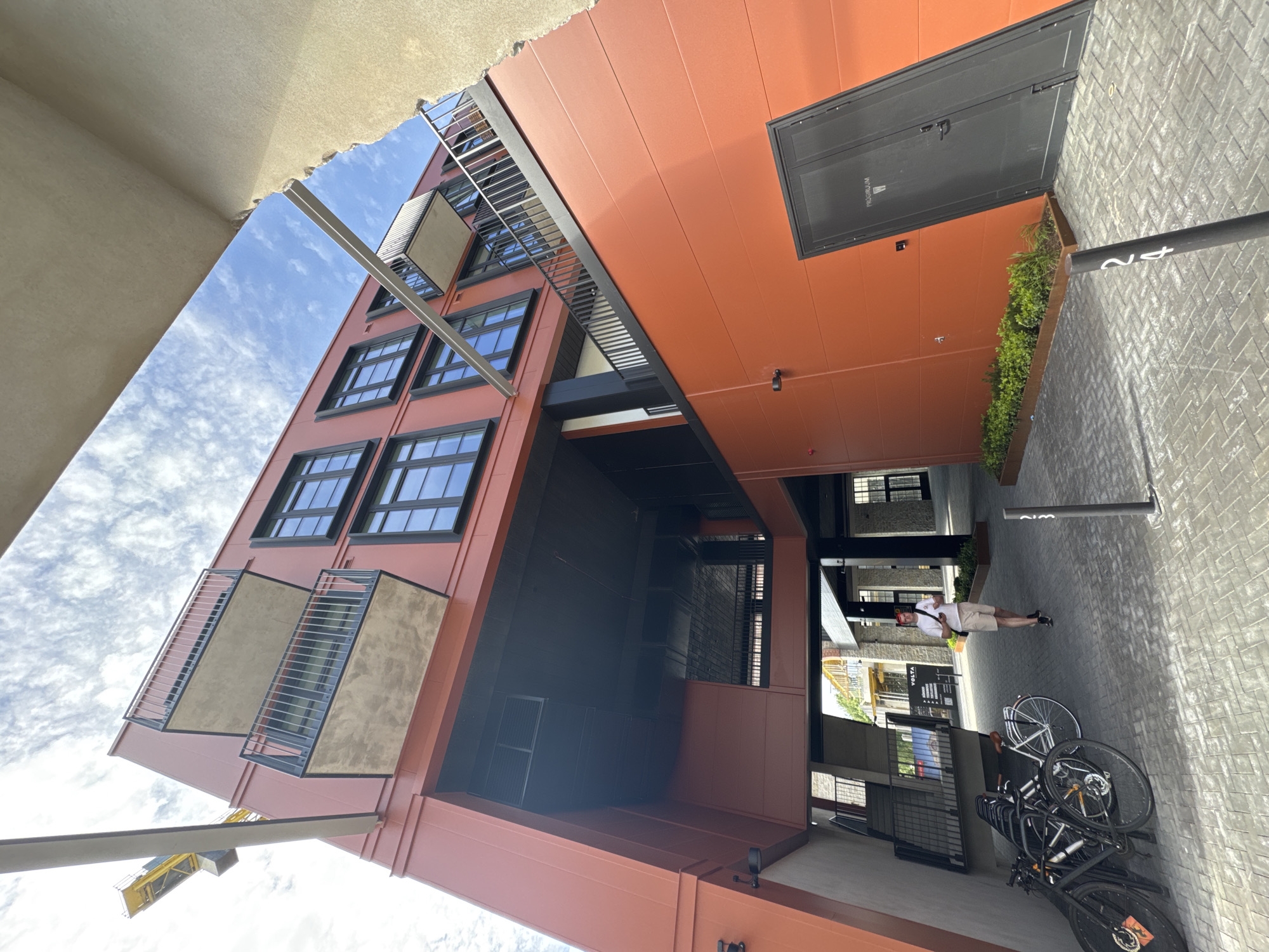 The image depicts an outdoor scene in Tallinn, Estonia, likely in a modern urban development. The focal point is a contemporary building with an orange facade, featuring multiple floors with dark-framed windows and balconies with metal railings. A portion of a yellow construction crane is visible in the upper left corner, suggesting ongoing development or recent construction. In the foreground, a person wearing a white t-shirt and beige shorts walks away from the camera on a cobblestone pathway. To the left, several bicycles are parked. The environment includes architectural elements such as covered walkways, planters with greenery, and a dark gray door with a sign indicating a restroom. The sky is blue with scattered clouds.