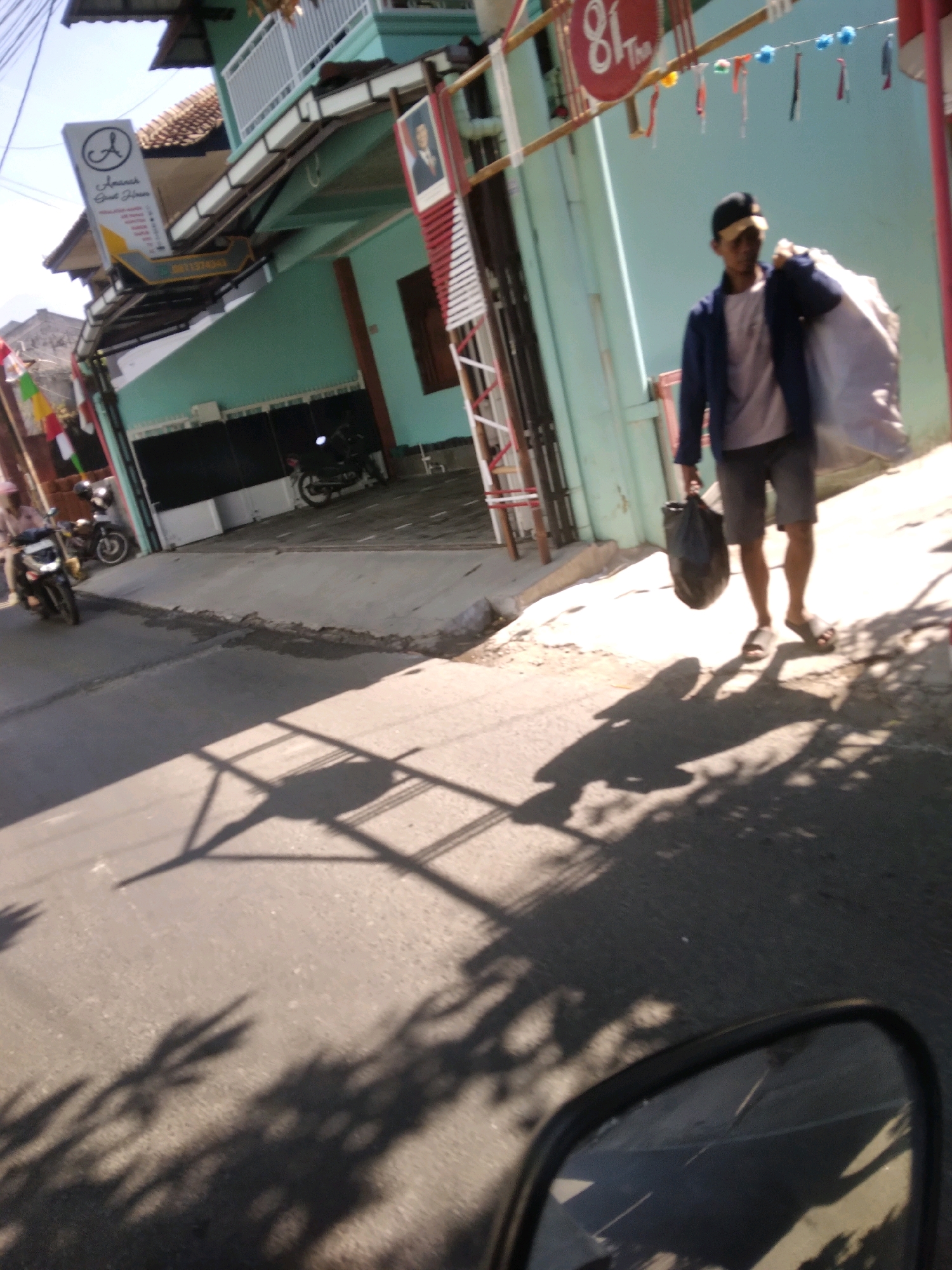 In Grendeng, Indonesia, a man walks down a paved street, carrying a large bag over his shoulder and a plastic bag in his hand. He is wearing a dark jacket, a light-colored t-shirt, dark shorts, a black baseball cap, and sandals. The street is lined with buildings. One building on the left has a sign with an "A" logo and text in Indonesian. On the right, a light blue building has a sign with "81 The" on it. Several motorcycles are parked in front of the buildings. The sun casts long shadows across the street, suggesting it is either morning or afternoon.