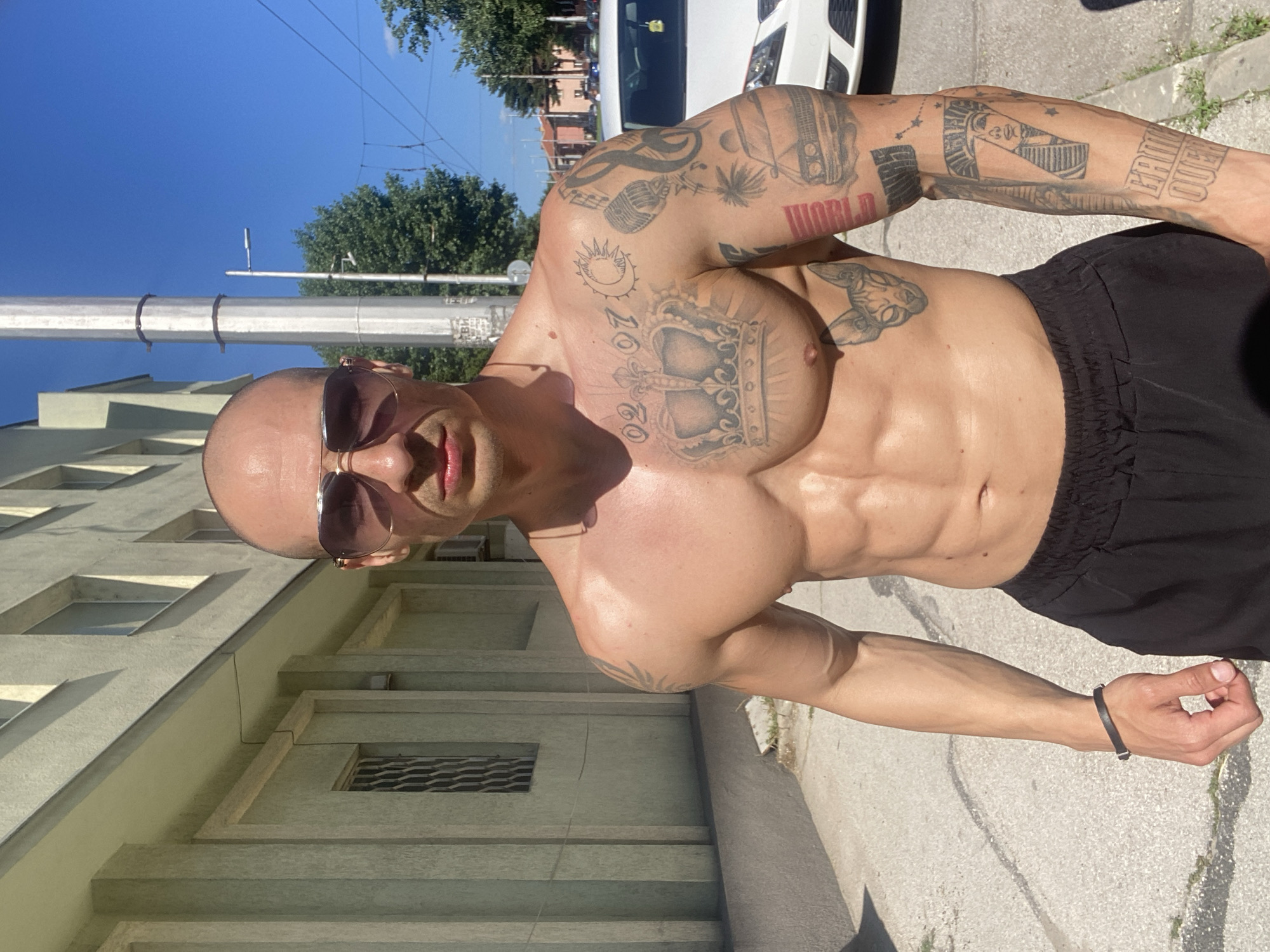 A muscular, bald man with tattoos is standing shirtless outdoors in front of a building. He is wearing sunglasses and black shorts. The tattoos are visible on his arms, chest, and torso. The background includes buildings, trees, a light pole, a white van, and a blue sky. The photo was taken in Pleven, Bulgaria.