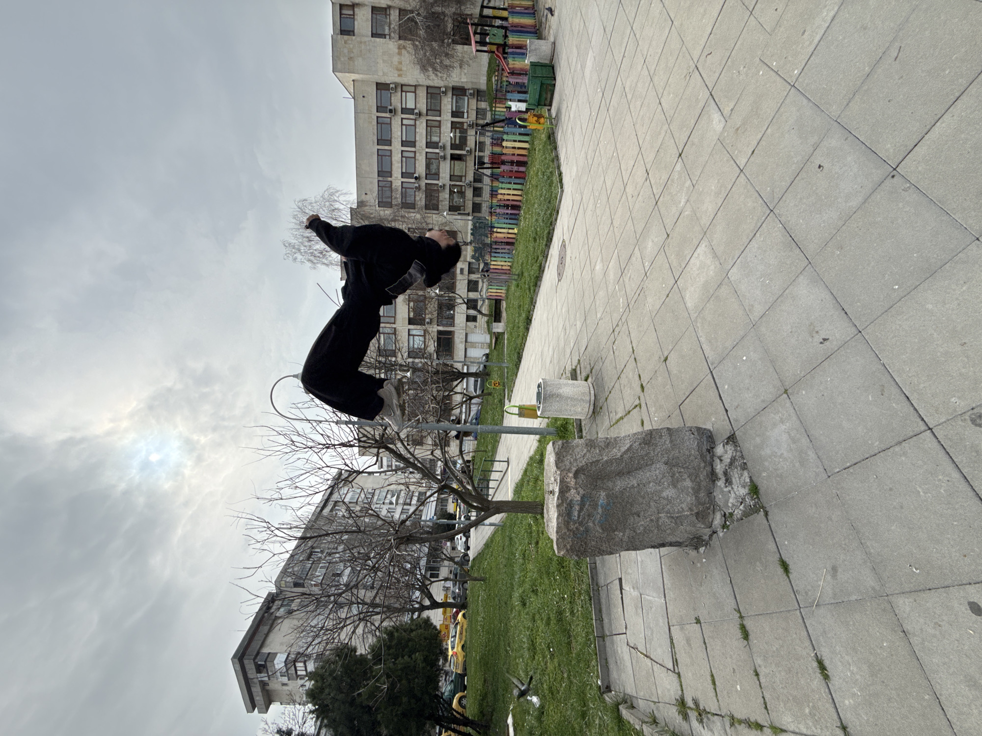 A person in mid-air performing a backflip, dressed in black clothing and white sneakers. The background includes a city park landscape, with a building visible in the backdrop, indicating the location is likely in an urban area. Buildings in the backdrop indicate the location of Shumen, Bulgaria, with playground sets and urban architecture visible. The person is positioned over a grassy area alongside a stone structure and a small tree. The scene is shot at an angle, possibly tilting the perspective of the urban environment. The sky is overcast, suggesting it's daytime.