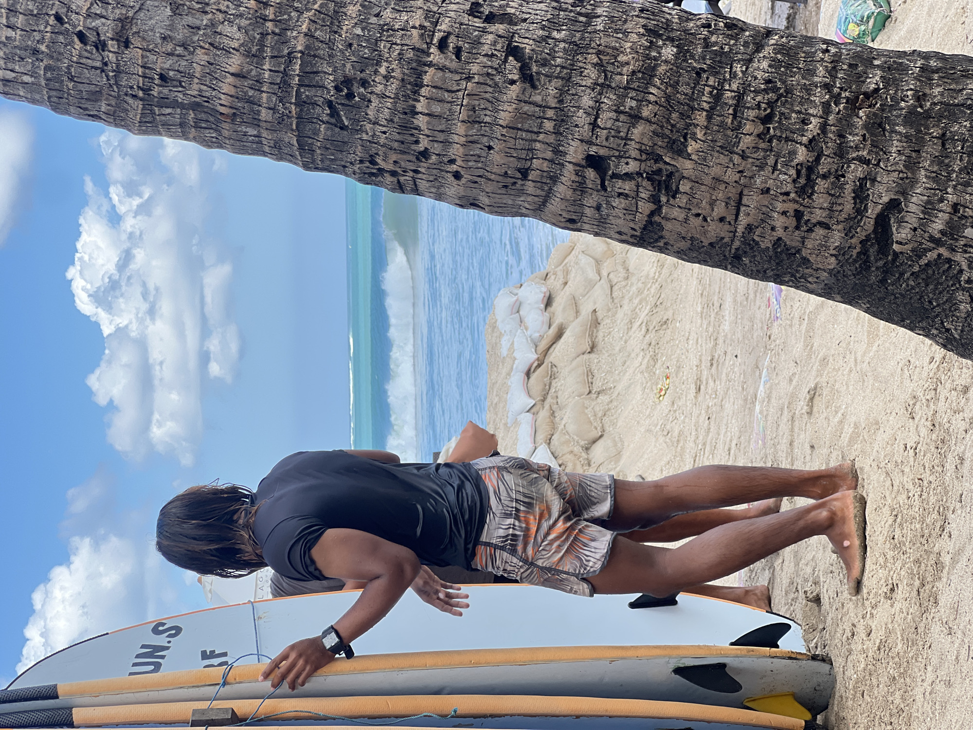 In Kuta, Indonesia, on a sandy beach with a backdrop of the ocean and a blue sky with scattered clouds, a man with wet, dark hair stands facing away from the camera. He is wearing a black t-shirt and patterned swim shorts. Next to him are two surfboards, one with orange accents and a white surface, and another that is partially visible. The man is holding the surfboard on the left. Behind him, the ocean has moderate waves. To the right of the frame, the rough, textured bark of a palm tree trunk is prominent, partially obscuring the view of the beach and ocean. Sandbags are piled up on the beach in the mid-ground.