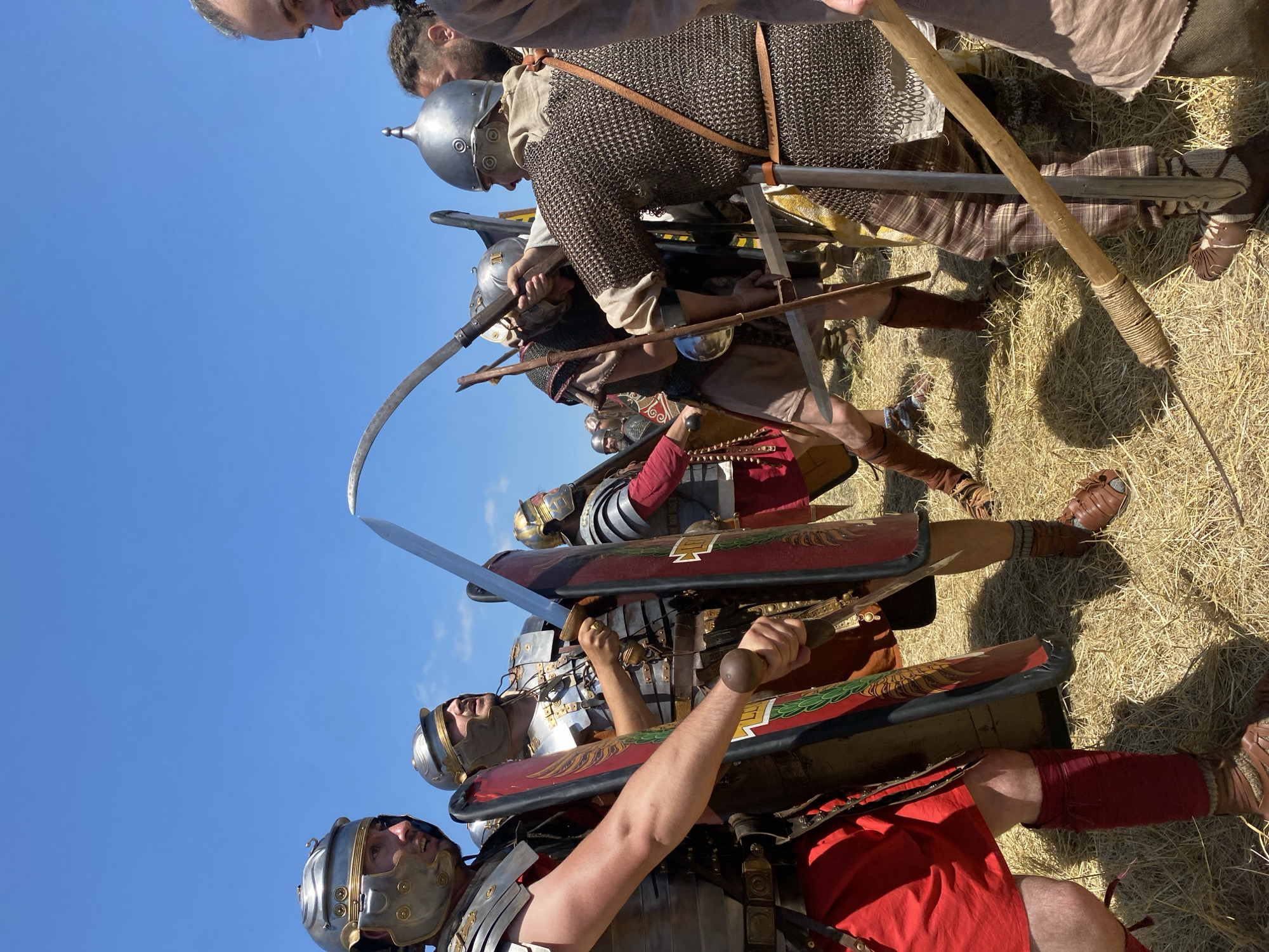 A wide-angle, eye-level photograph captures a historical reenactment scene outdoors under a clear blue sky in Gigen, Bulgaria. The ground is covered with dry, golden straw or hay. The scene depicts a close-quarters combat engagement between individuals dressed in ancient military attire, illuminated by bright sunlight.
In the left foreground, a male participant, identified as a Roman soldier, wears a silver-colored helmet with brass trim, segmented plate armor (lorica segmentata), a red tunic, and brown leg wraps. He holds a large, rectangular red and green shield (scutum) on his left arm and a gladius-style short sword with a spherical pommel in his right hand, positioned defensively. His profile is visible, gazing towards the central action.
To his right, another Roman soldier, similarly armored with a helmet and lorica segmentata, holds a straight sword aloft and a scutum. He is partially obscured by the foreground soldier and appears to be actively engaging an opponent.
In the center-right, a warrior in chainmail (lorica hamata) and a dome-shaped helmet with a pointed top actively wields a large, curved sword (likely a falx or sica) with a wooden hilt, bringing it down towards an unseen opponent. His face, partially obscured, shows concentration. Another straight sword is visible intersecting with his curved weapon. A long wooden spear or javelin (pilum) rests vertically against his left leg.
Further to the right, a male observer or participant with a beard and braided hair is partially visible, wearing a light-colored tunic, watching the combat.
In the background, additional reenactors in various forms of Roman and potentially other period armor and clothing are visible, some forming a line, indicating a larger group presence. Shadows are visible on the ground beneath the participants, indicating a high sun.